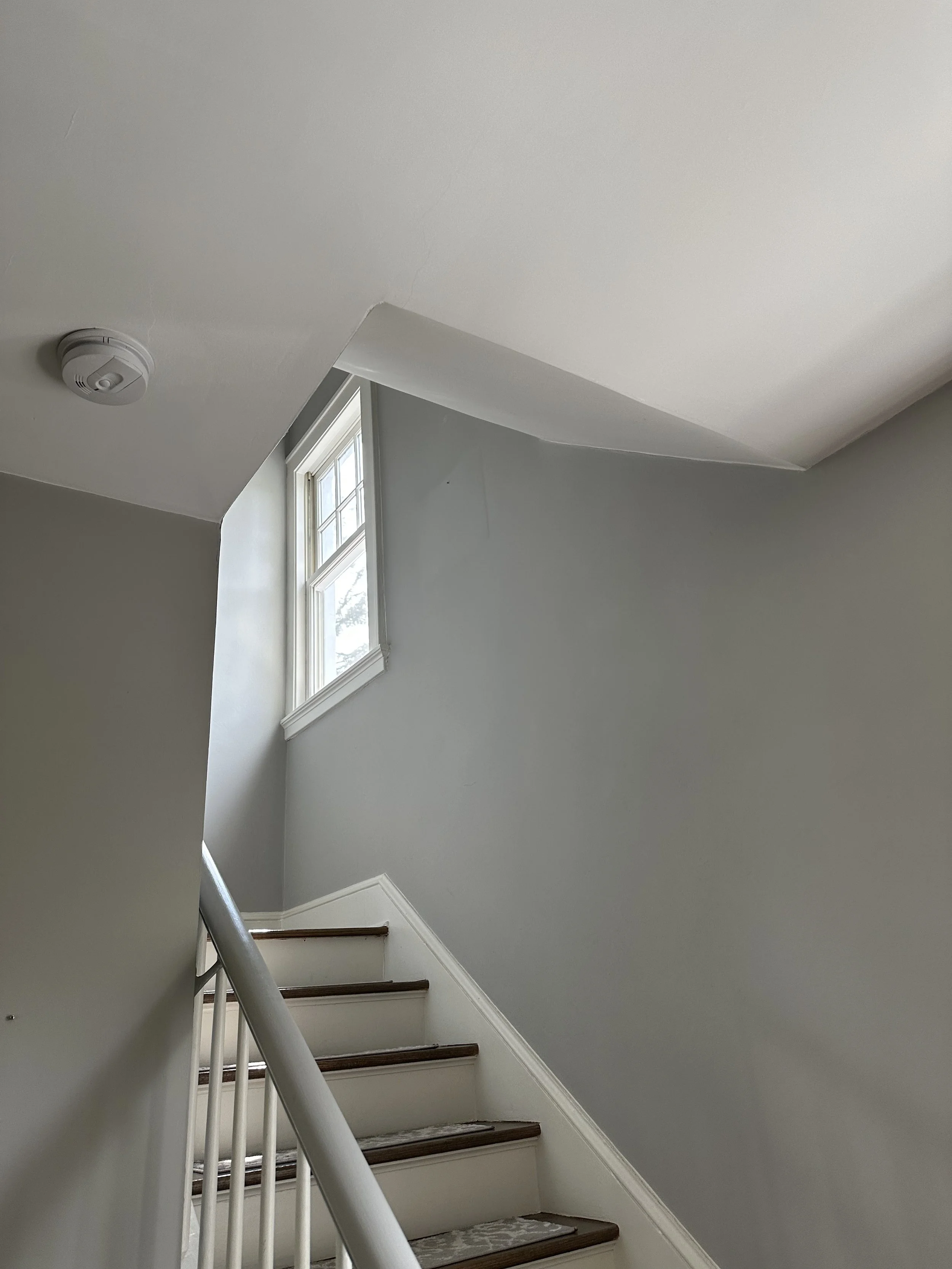 Interior view of a staircase with a window at the top, gray walls, wooden stairs, and a smoke detector on the ceiling.