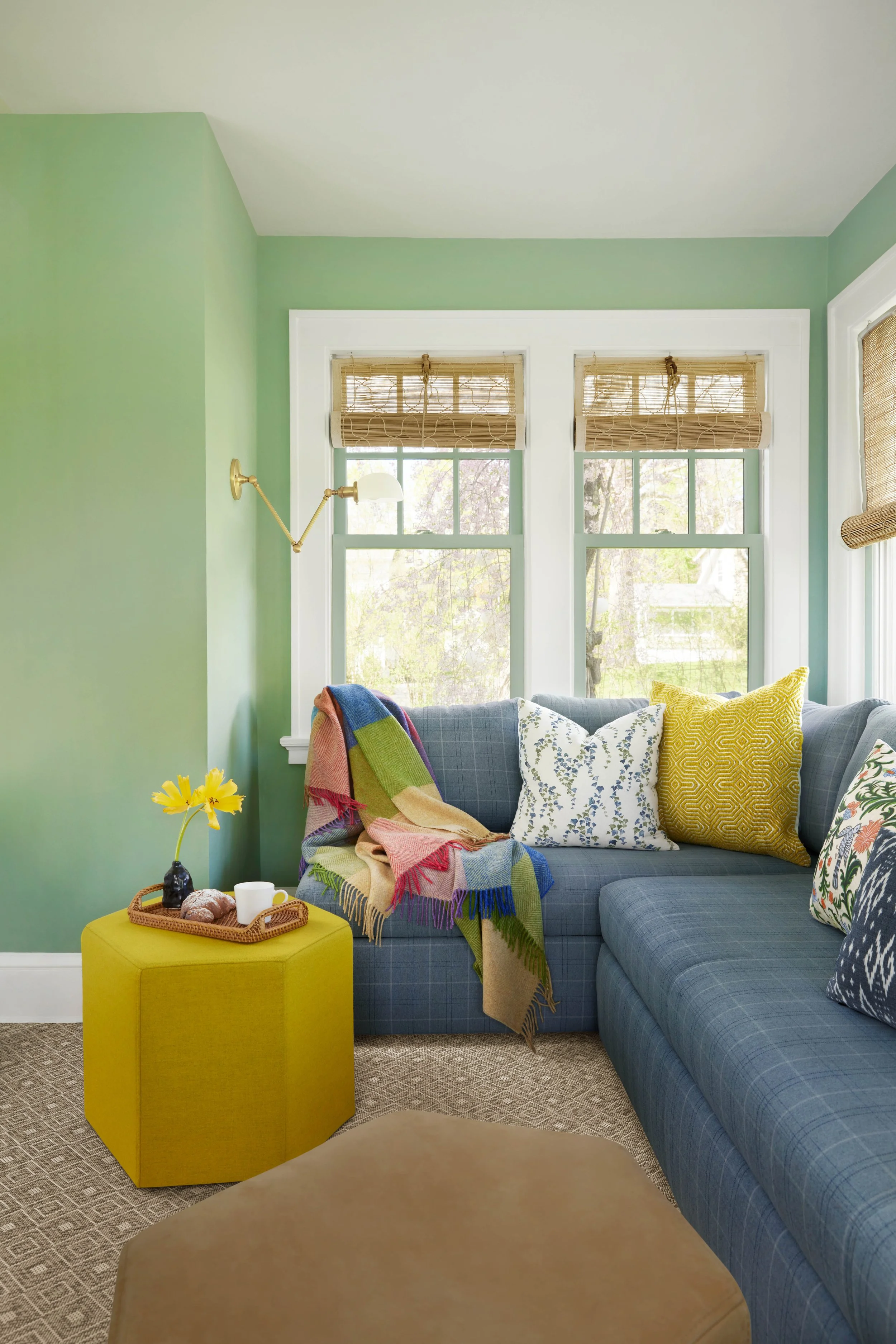 Cozy living room corner with a blue plaid sectional sofa, yellow and floral throw pillows, a yellow ottoman with a tray holding a cup, croissants, and a vase with a yellow flower. Green walls and large windows with bamboo shades.