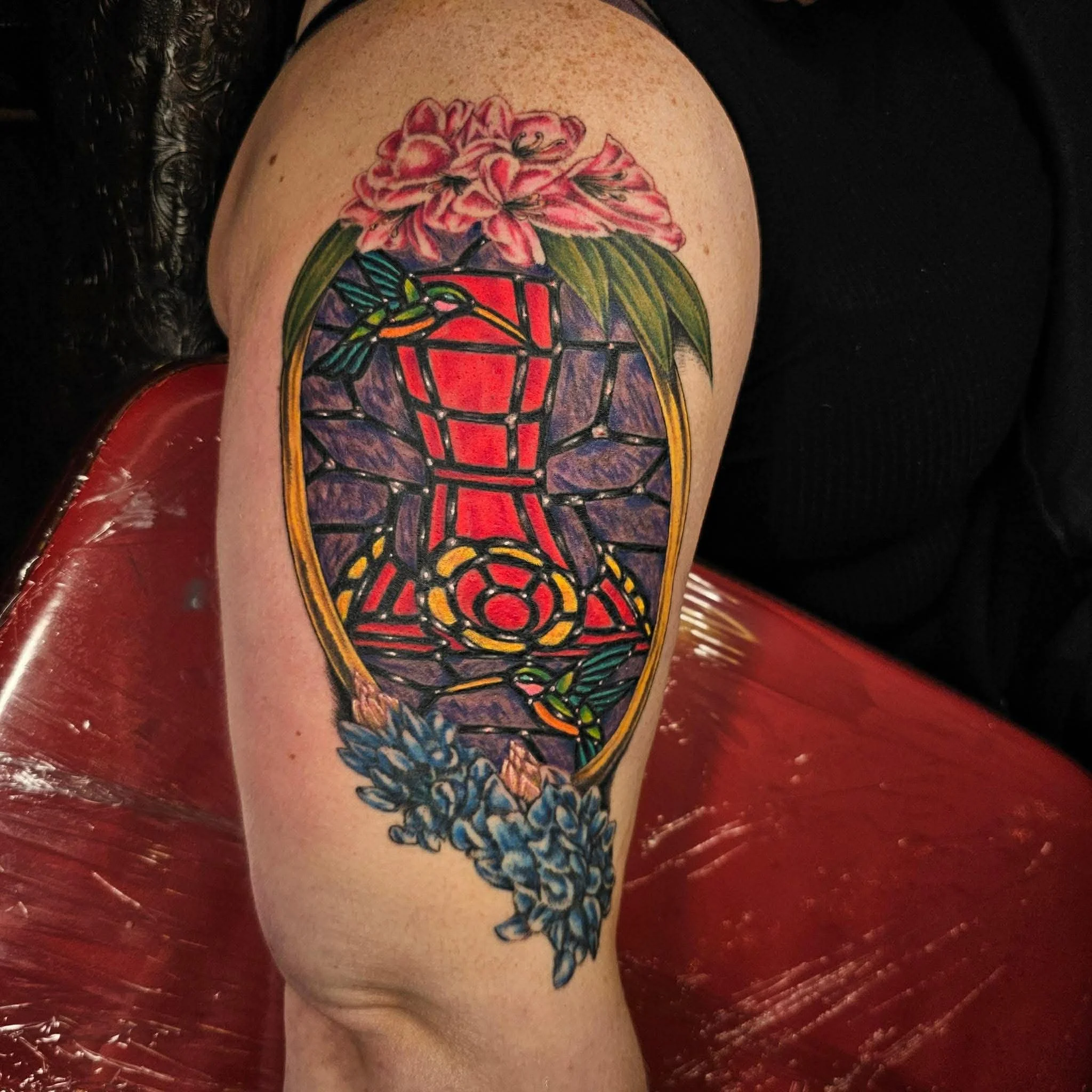 A tattoo of stained glass art with a humming bird feeder and humming birds; flowers surround the top and bottom edges.