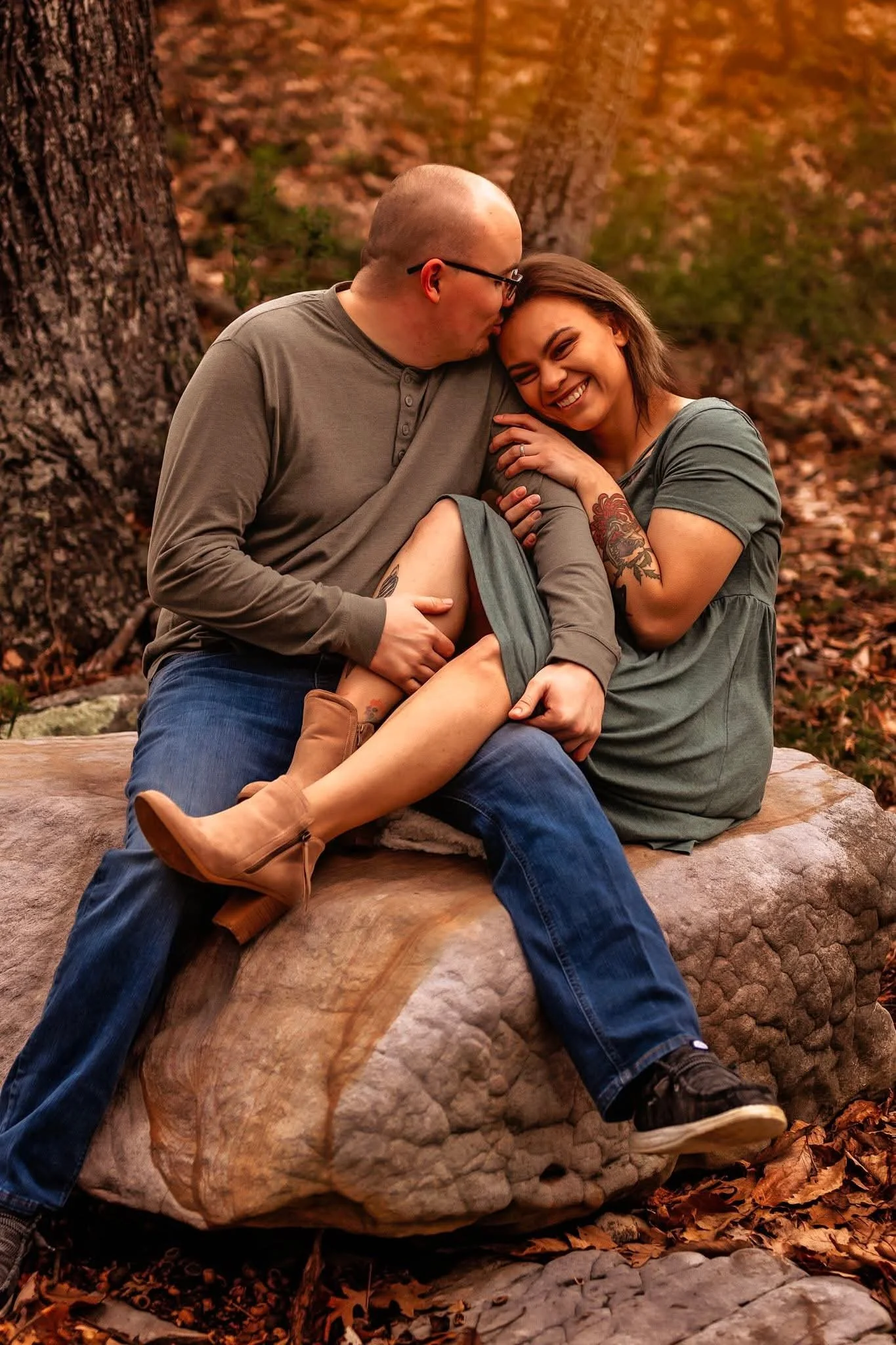 Sarah and her husband posing on a rock. She smiles and the camera while holding his arm. He kisses her on the forehead.
