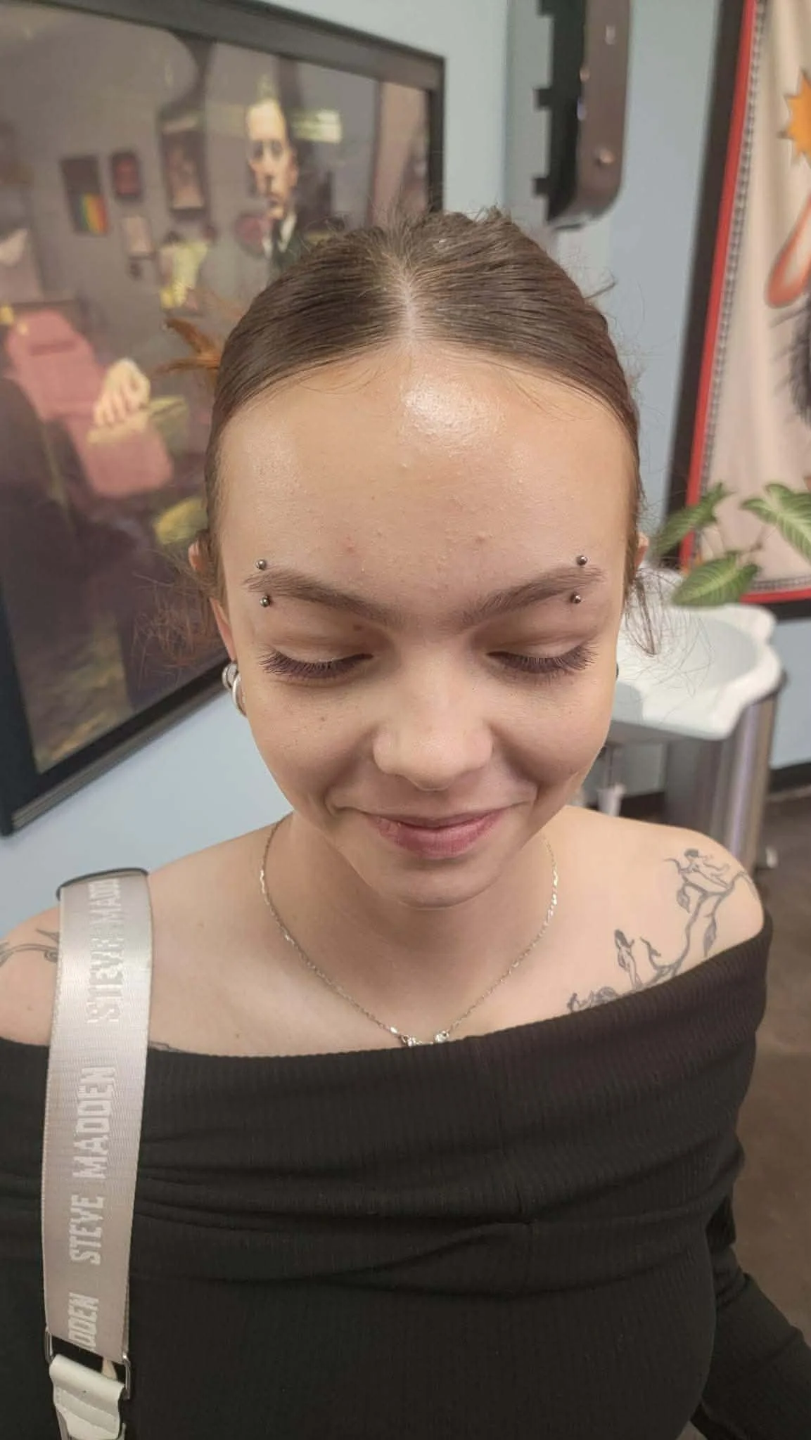 A woman smiles and looks down. She has two matching vertical eyebrow piercings.