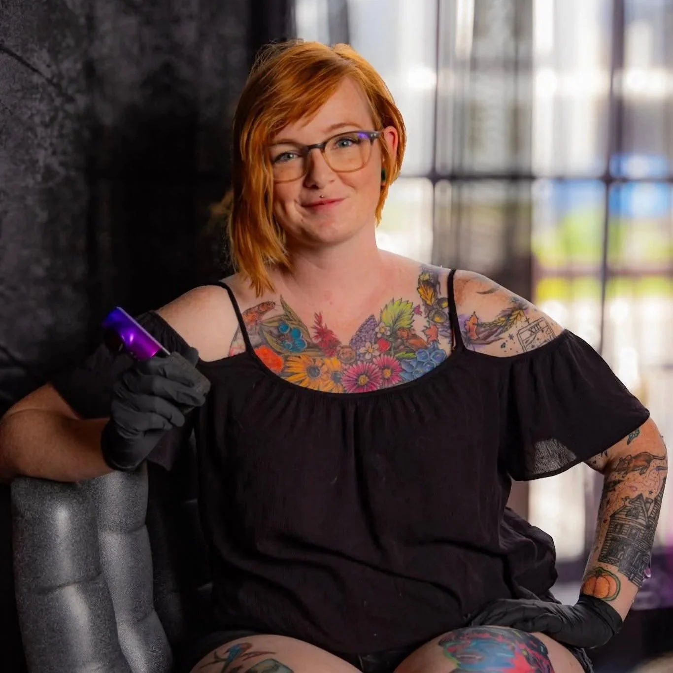 Mel sits in a chair holding a tattoo machine and smiling.