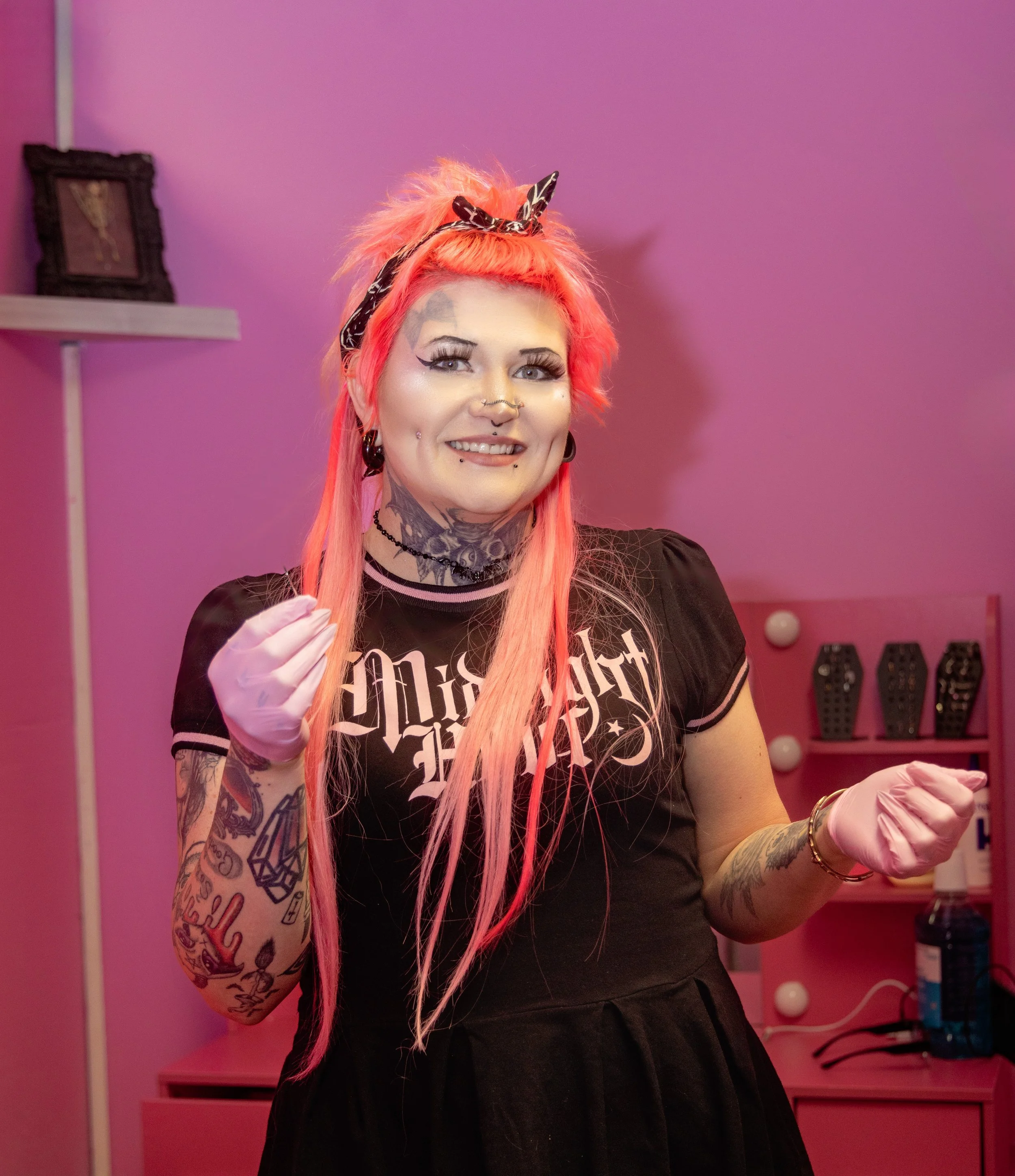 A photo of Hexed Aesthetic (Sheena) smiling in her piercing room. She is wearing gloves and holding a needle.