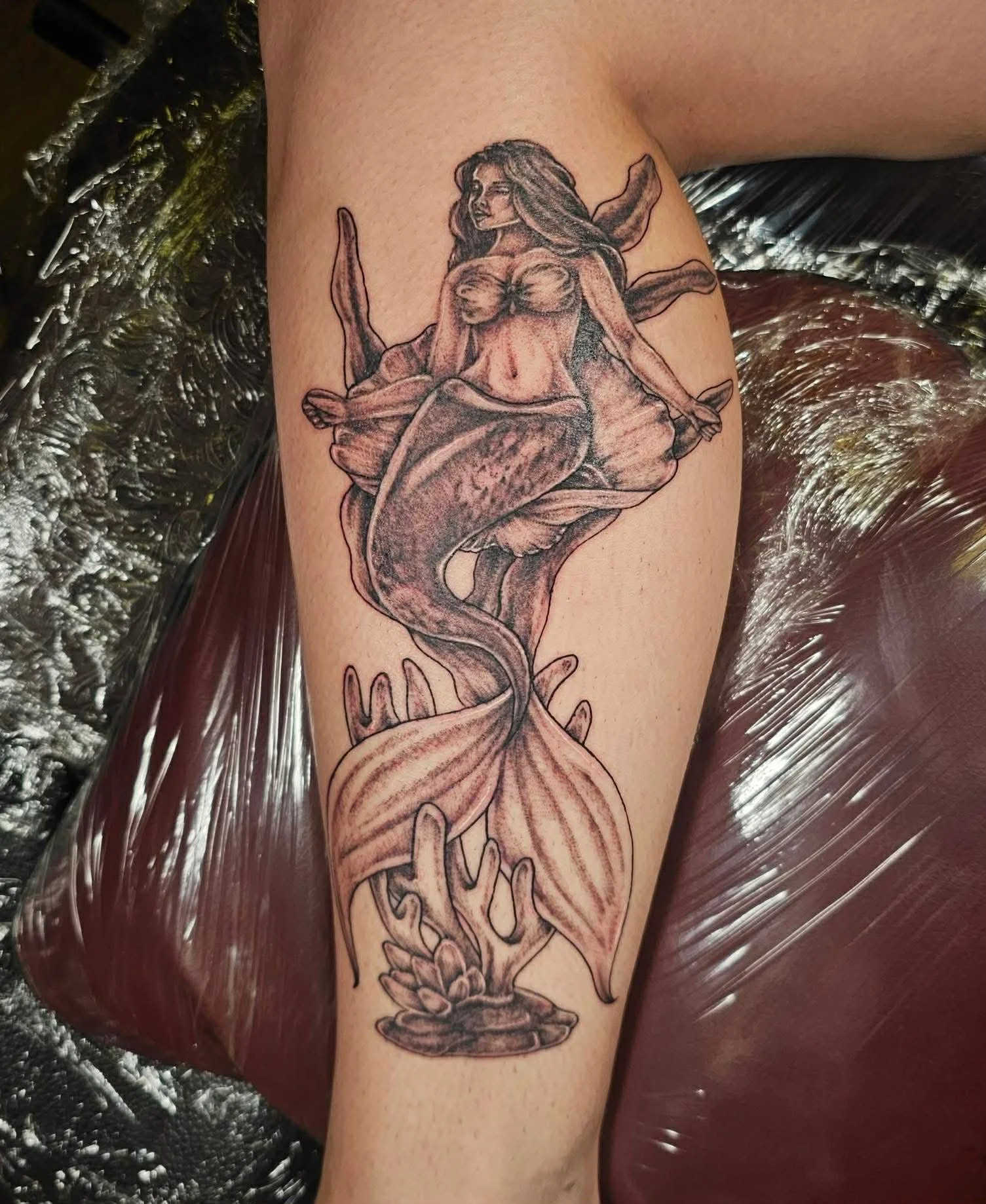 A greyscale tattoo of a mermaid siting on a large shell, surrounded by coral. 