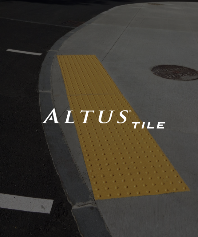 Close-up of a yellow tactile paving tile on a city sidewalk at a corner, with a dark street and a manhole cover nearby.