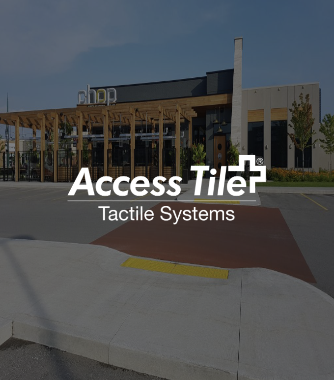Exterior of a commercial building with a wooden awning and trees, with the logo 'Access Tiler Tactile Systems' and a tactile warning strip on the sidewalk in front.
