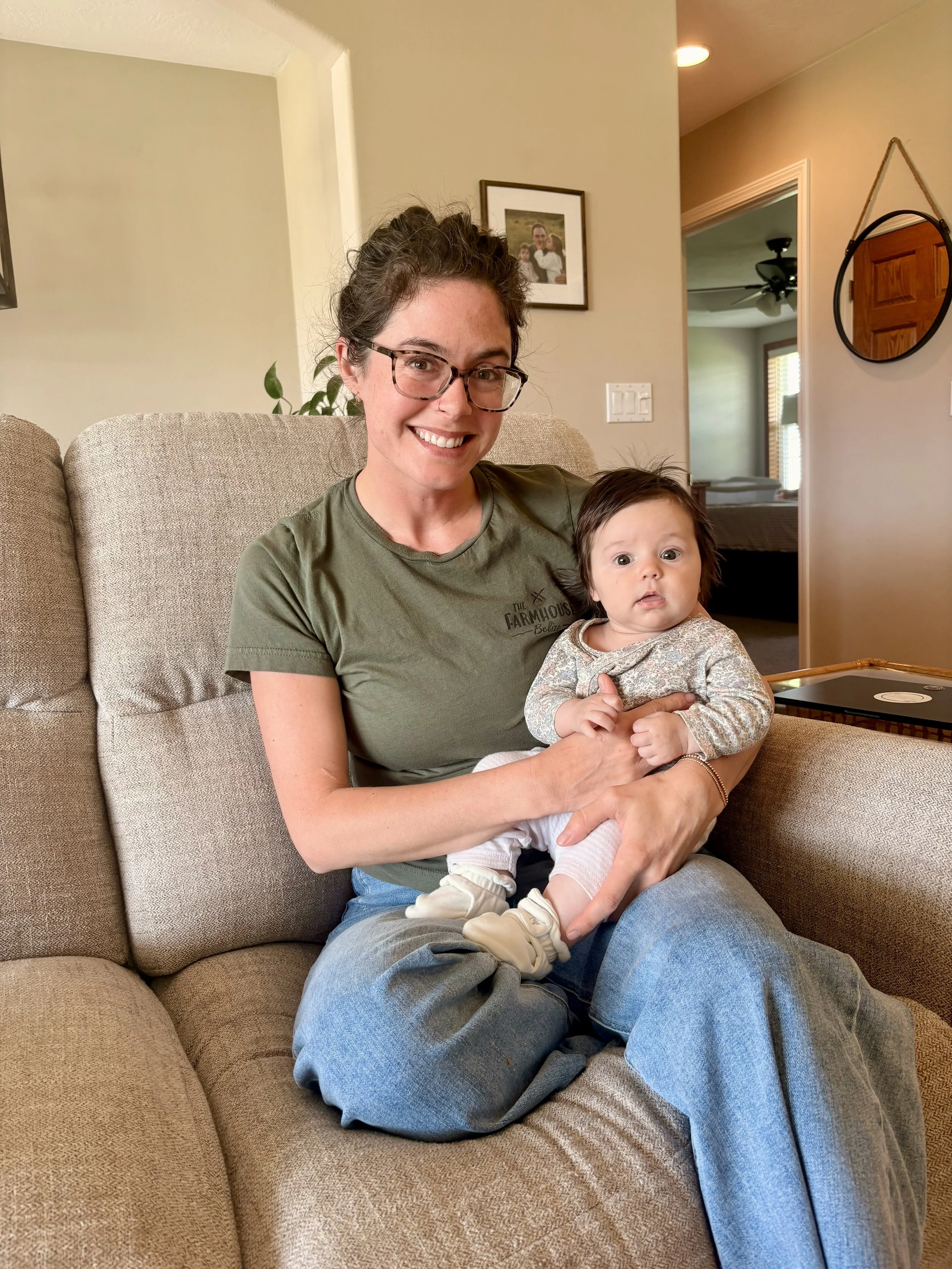 A woman with glasses and a green shirt smiling and holding a young girl with brown hair and big eyes sitting on her lap on a beige sofa in a living room.
