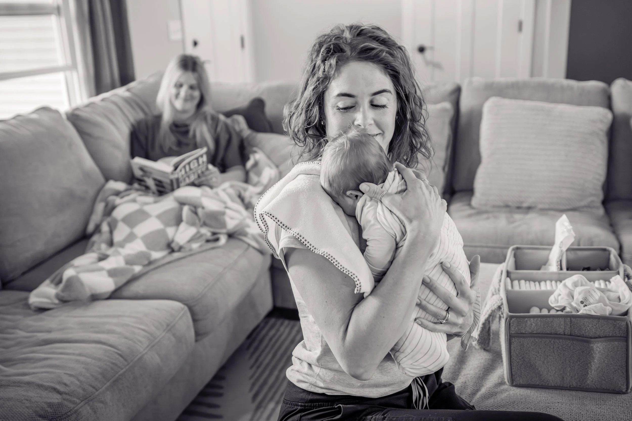 A woman holding a baby close to her chest while sitting on the floor in a living room, with an older woman sitting on a couch reading a book in the background.