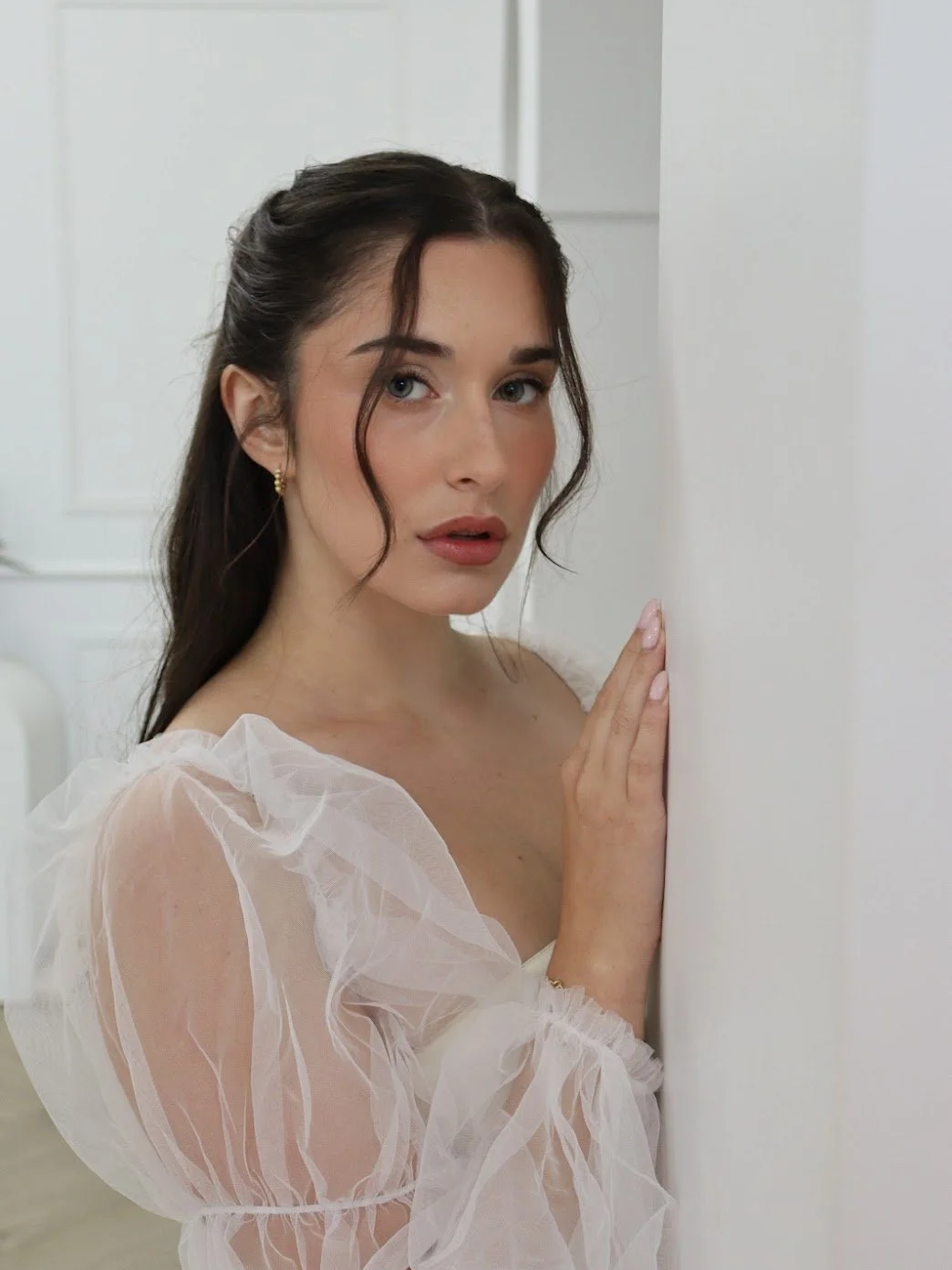 A woman with long dark hair and blue eyes, wearing a sheer, ruffled white blouse, touching a white wall and looking at the camera.