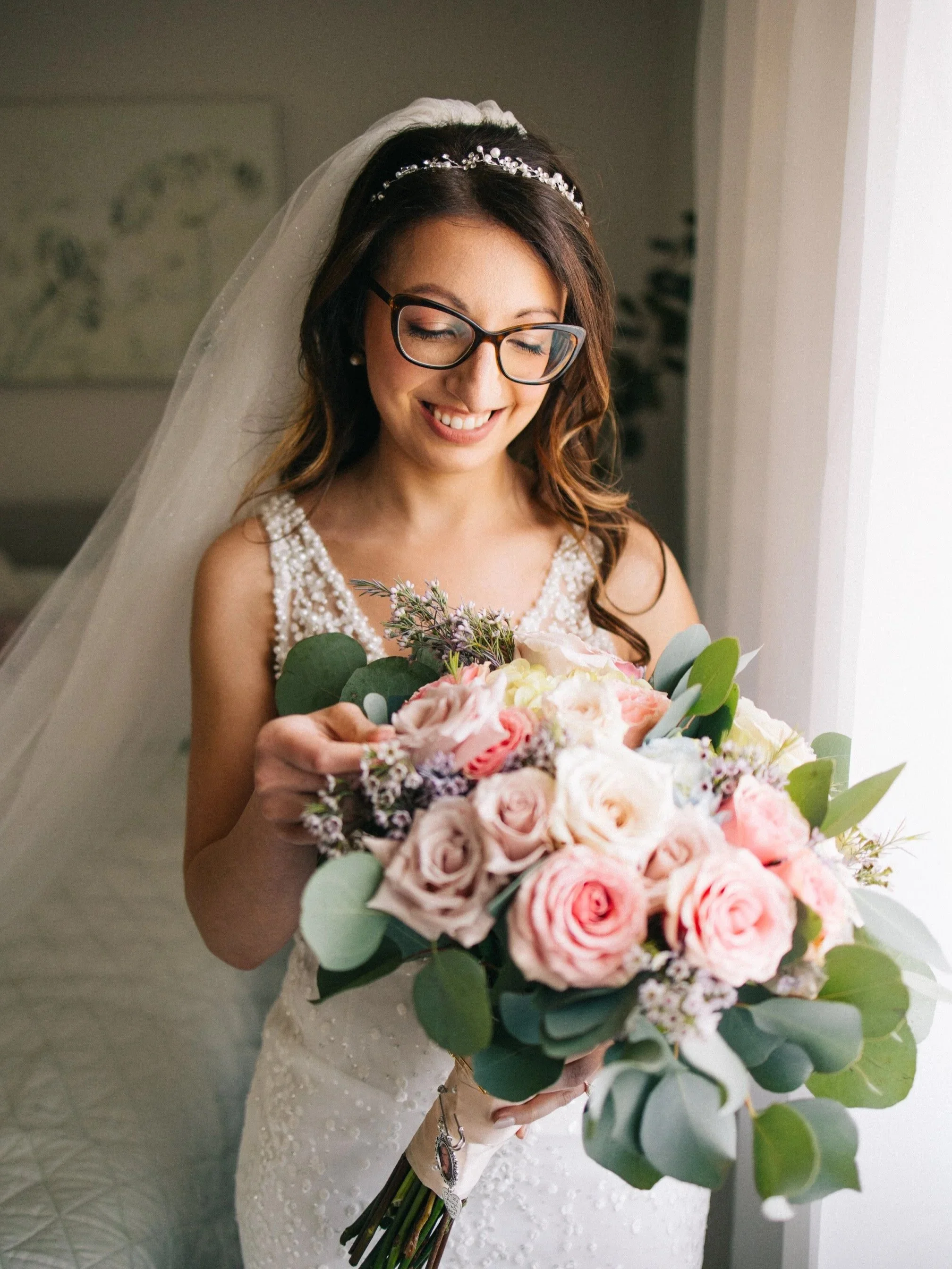 A bride with glasses, wearing a wedding dress and veil, holding a large bouquet of pink and white roses with greenery, standing near a window.
