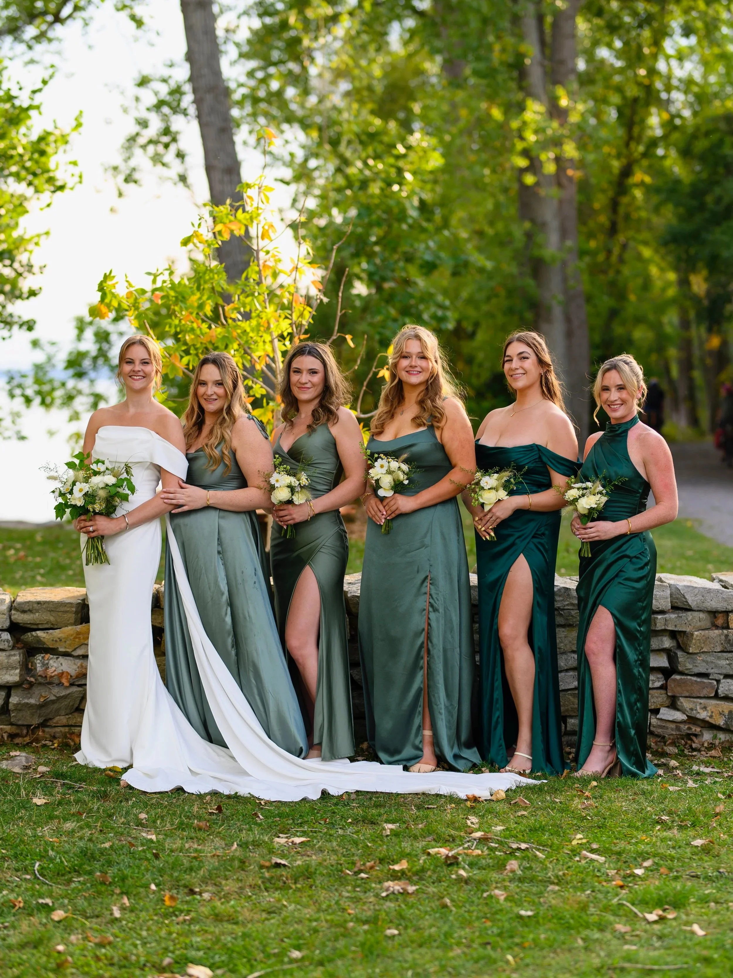 Group of six women at an outdoor wedding, wearing elegant dresses in shades of green and white, holding white floral bouquets, standing on a grassy area with trees in the background.