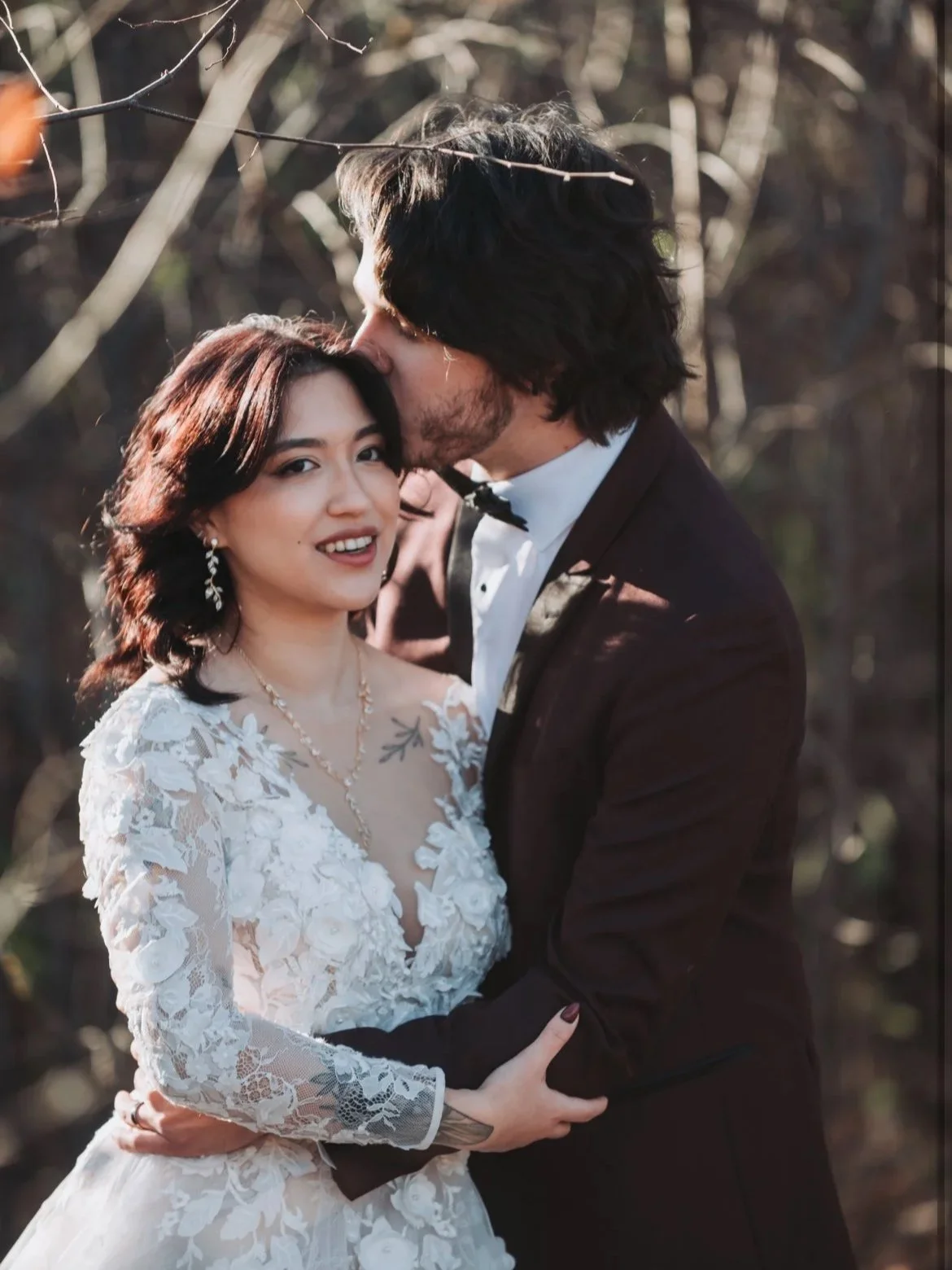 A bride and groom in wedding attire outdoors; the groom kisses the bride's forehead, the bride smiling at camera.