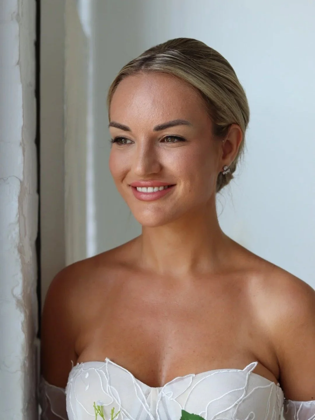 A bride with blonde hair styled in an elegant updo, wearing a strapless white dress with floral embroidery, standing near a window and smiling.