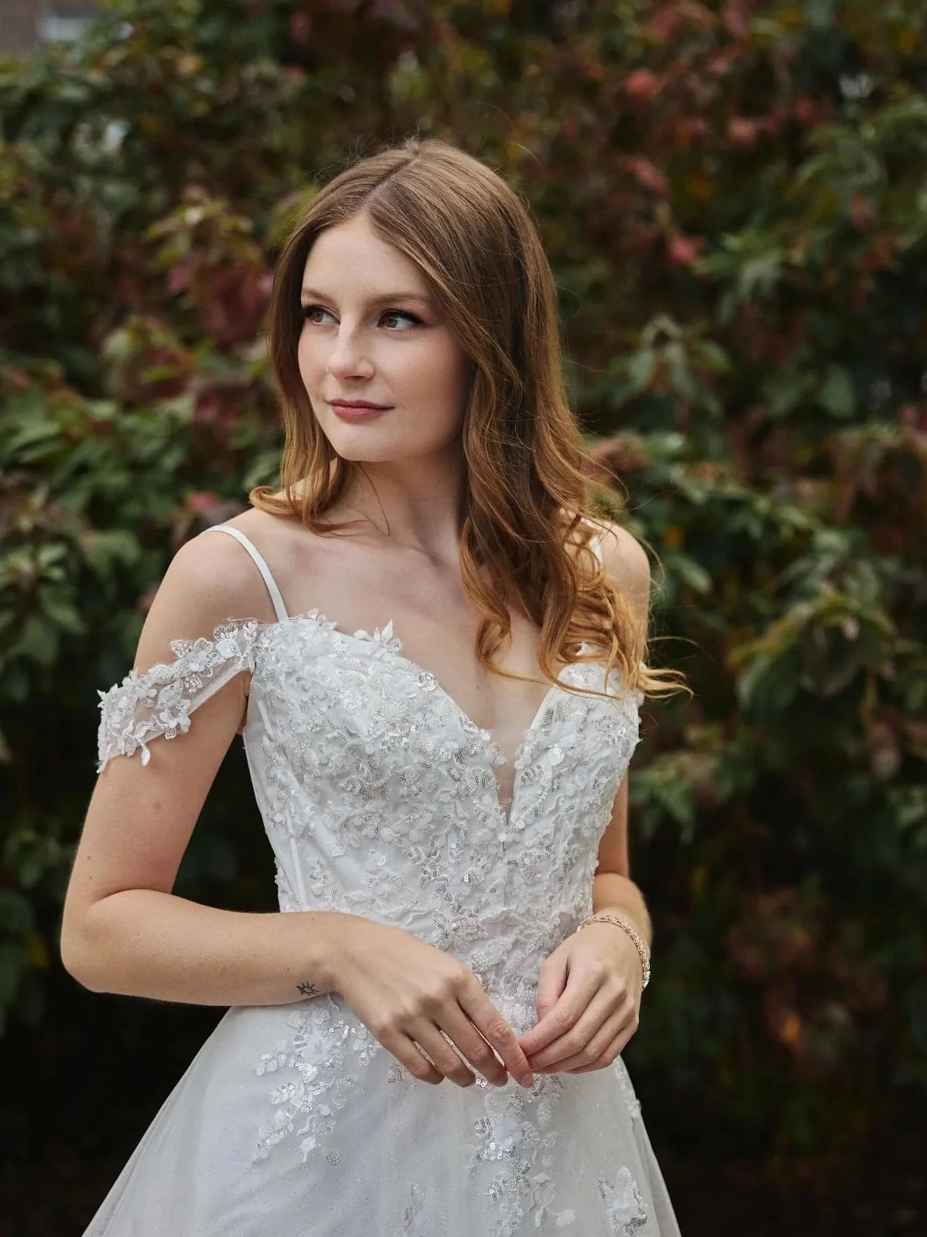 A bride in a white wedding dress with lace and floral details stands outdoors with a background of green and autumn-colored foliage.