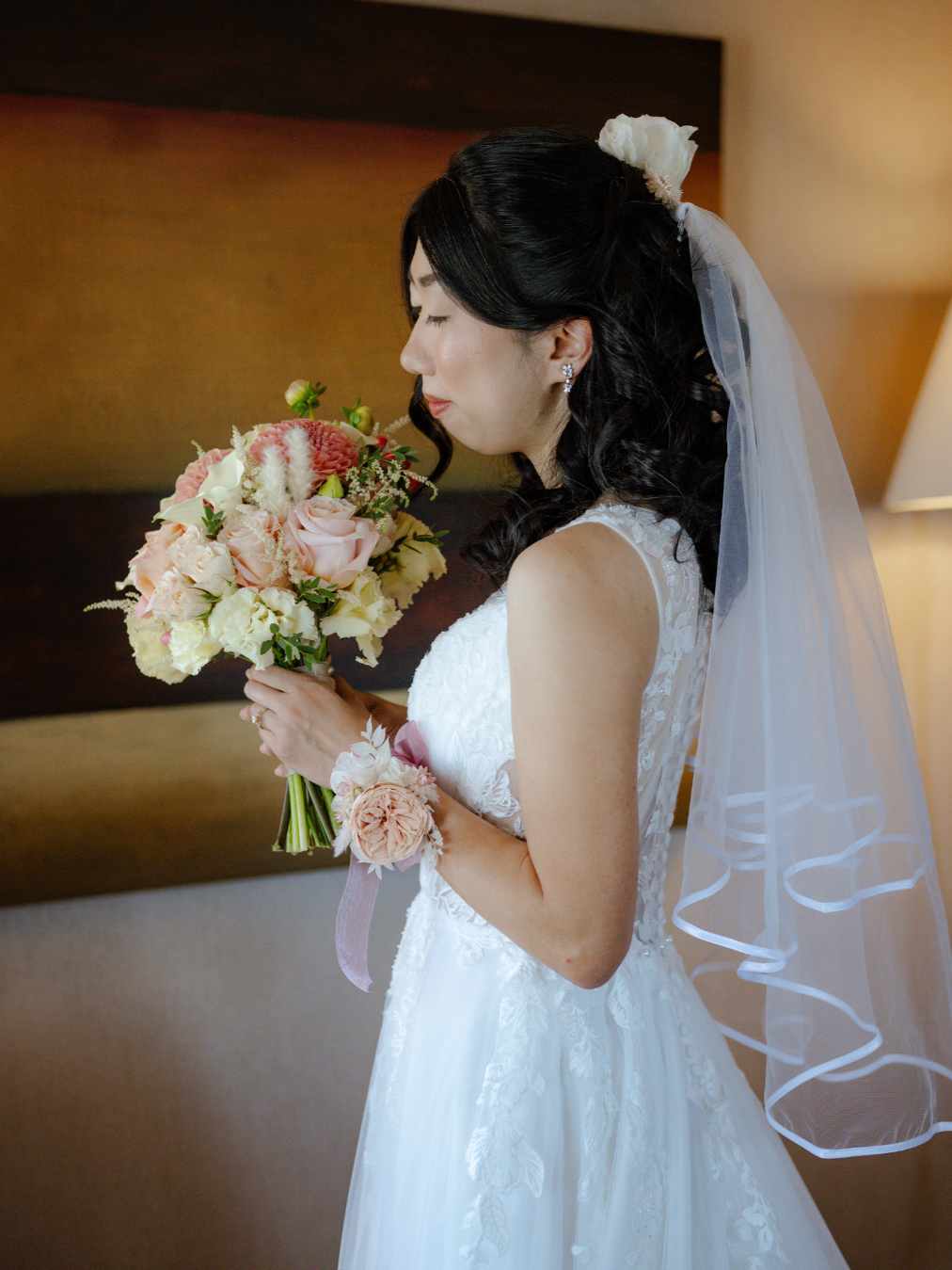 A bride in a white wedding dress holding a bouquet of pink and white flowers, with her eyes closed and head tilted slightly downward.