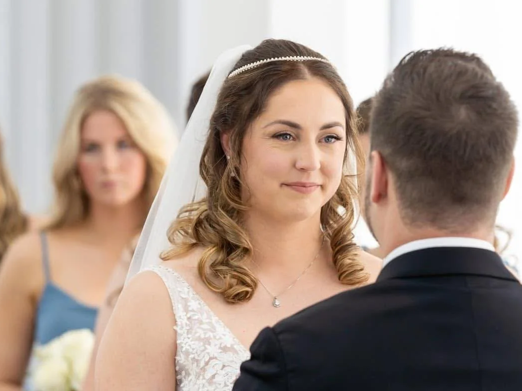 A bride and groom facing each other during their wedding ceremony, with a woman in the background holding a bouquet.