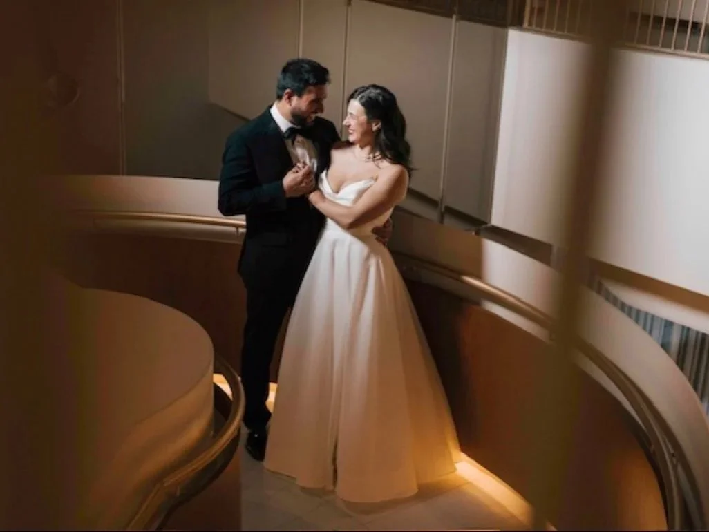 A couple dressed in wedding attire sharing a dance on a staircase, smiling and looking at each other.