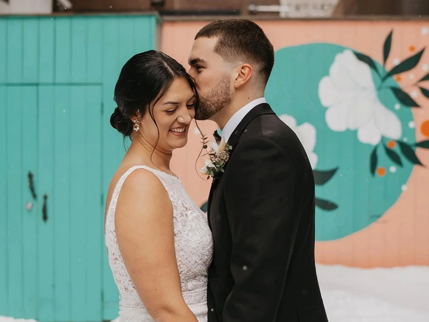 A bride and groom sharing a kiss, with the groom giving a gentle kiss on the bride's forehead, smiling, against a colorful mural background.