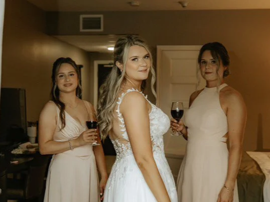 A bride and two bridesmaids in formal dresses at an indoor gathering, holding glasses of red wine.