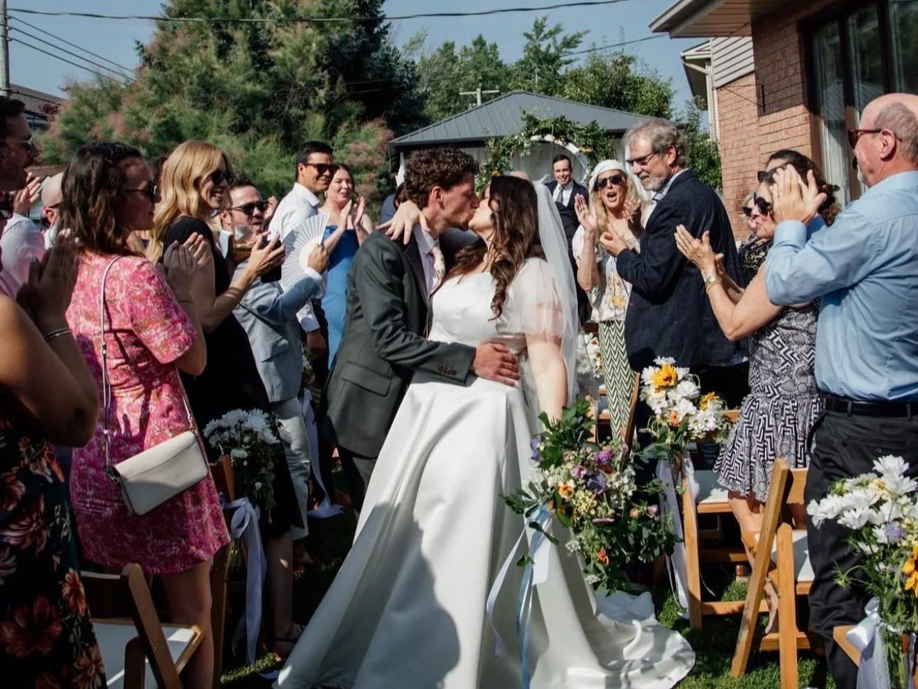 A wedding celebration with a bride and groom kissing in front of family and friends outdoors, with floral decorations and chairs.