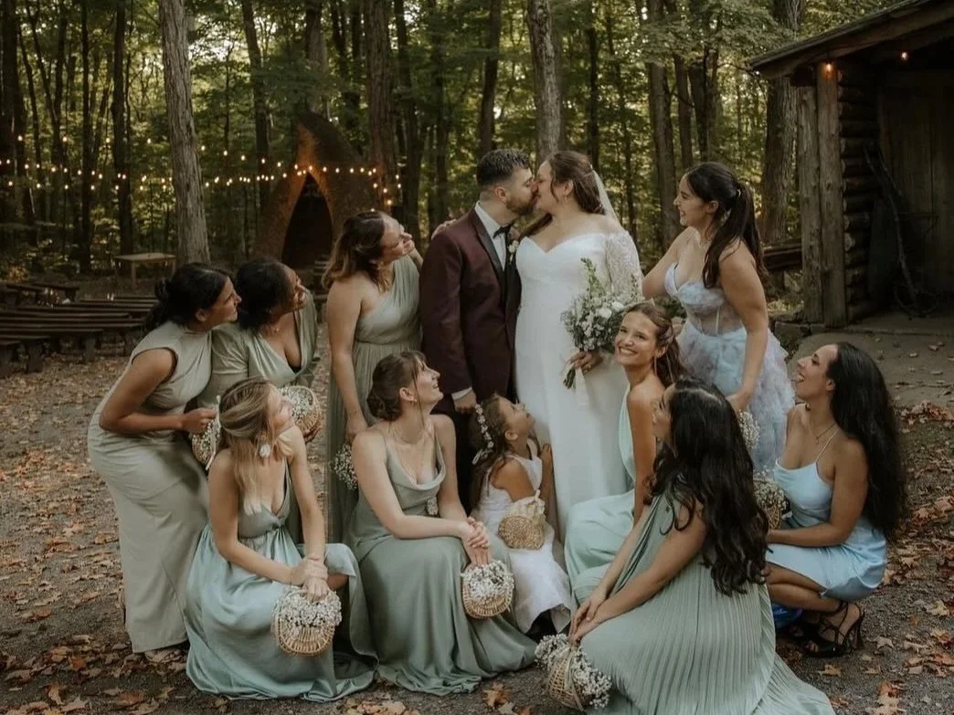 A wedding scene in a wooded outdoor setting with a bride and groom kissing surrounded by bridesmaids and flower girls, with string lights and a small cabin in the background.