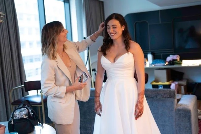 A woman in a wedding dress smiling while another woman adjusts her hair in a hotel room.