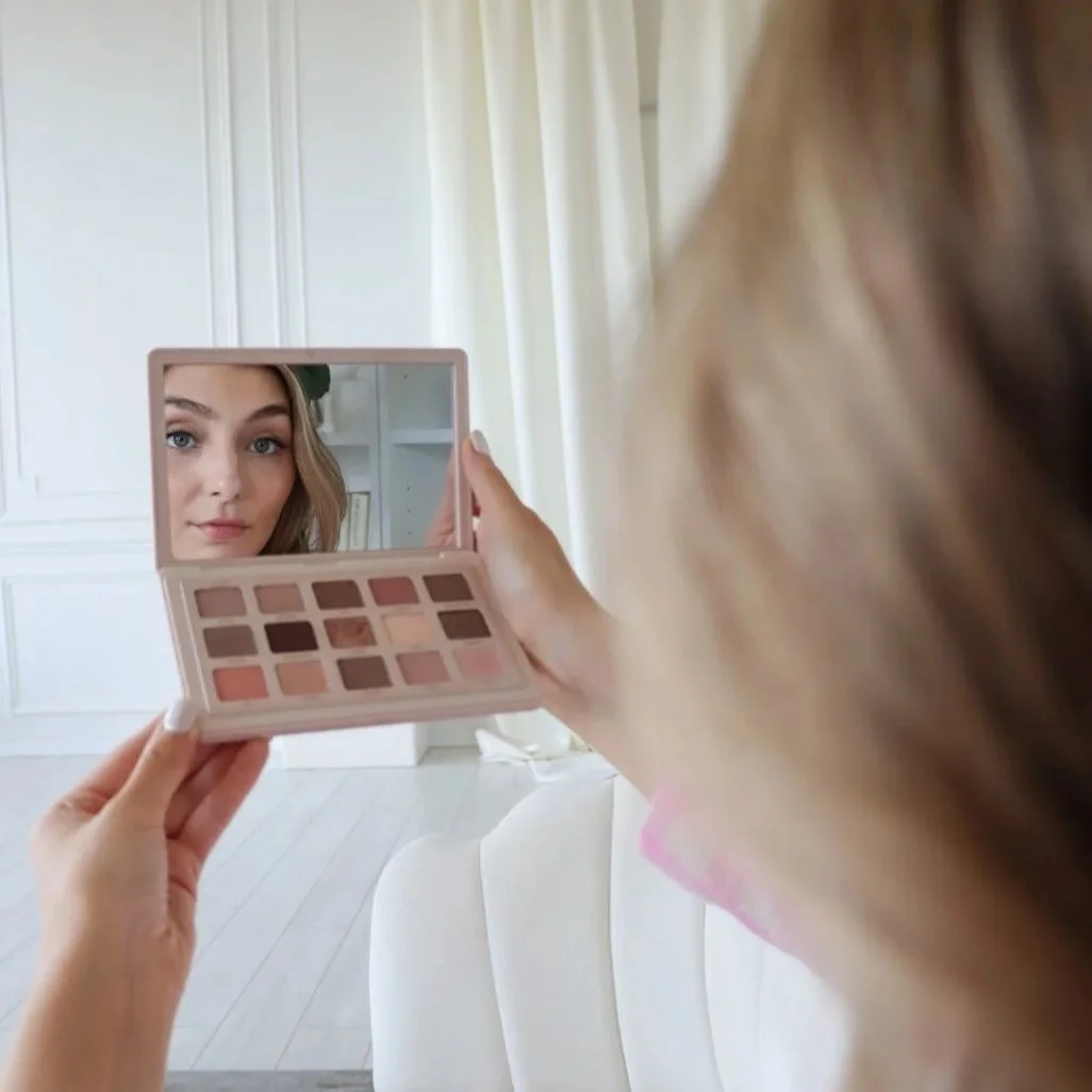 A woman is holding a makeup palette and a small mirror, applying makeup while looking at her reflection on the mirror.