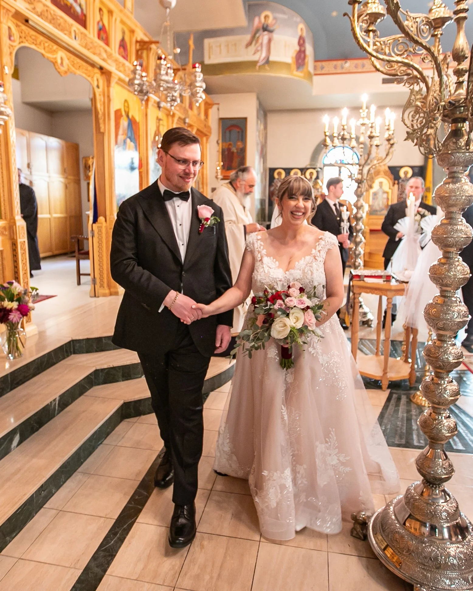A bride and groom holding hands inside a church during their wedding ceremony. The bride is holding a bouquet of pink and white flowers and is wearing a white lace wedding dress, while the groom is dressed in a black tuxedo with a bowtie. The church has ornate gold decor and religious icons in the background.