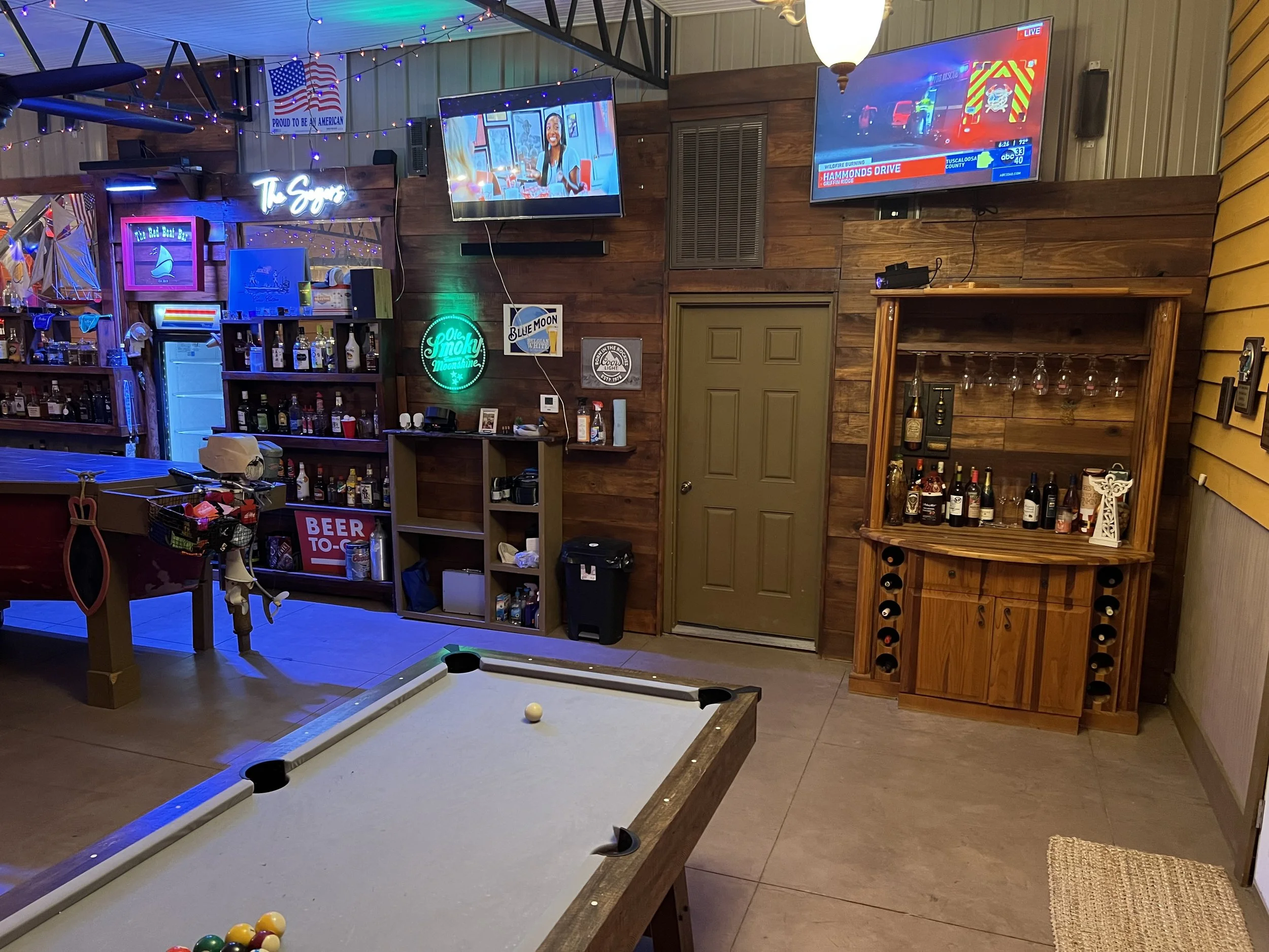 Indoor recreational space featuring a pool table with balls, a bar with bottles, two wall-mounted TVs, neon signs, and various decorations including a flag and string lights.