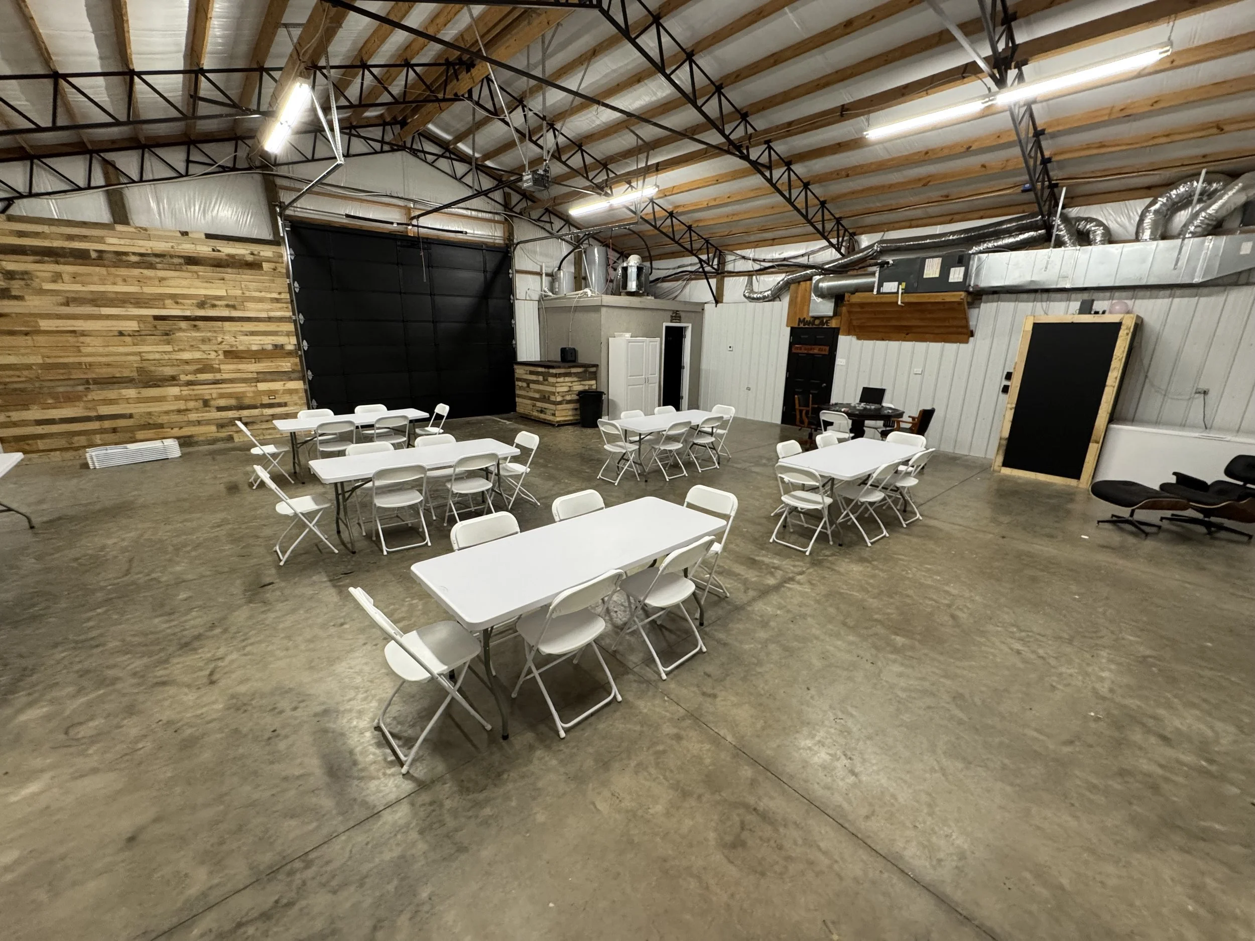 An indoor event space with several white tables and foldable chairs arranged for a gathering. The room has high ceilings with exposed wooden beams and ductwork, a black garage door on one wall, a wooden accent wall, and a black chalkboard or bulletin