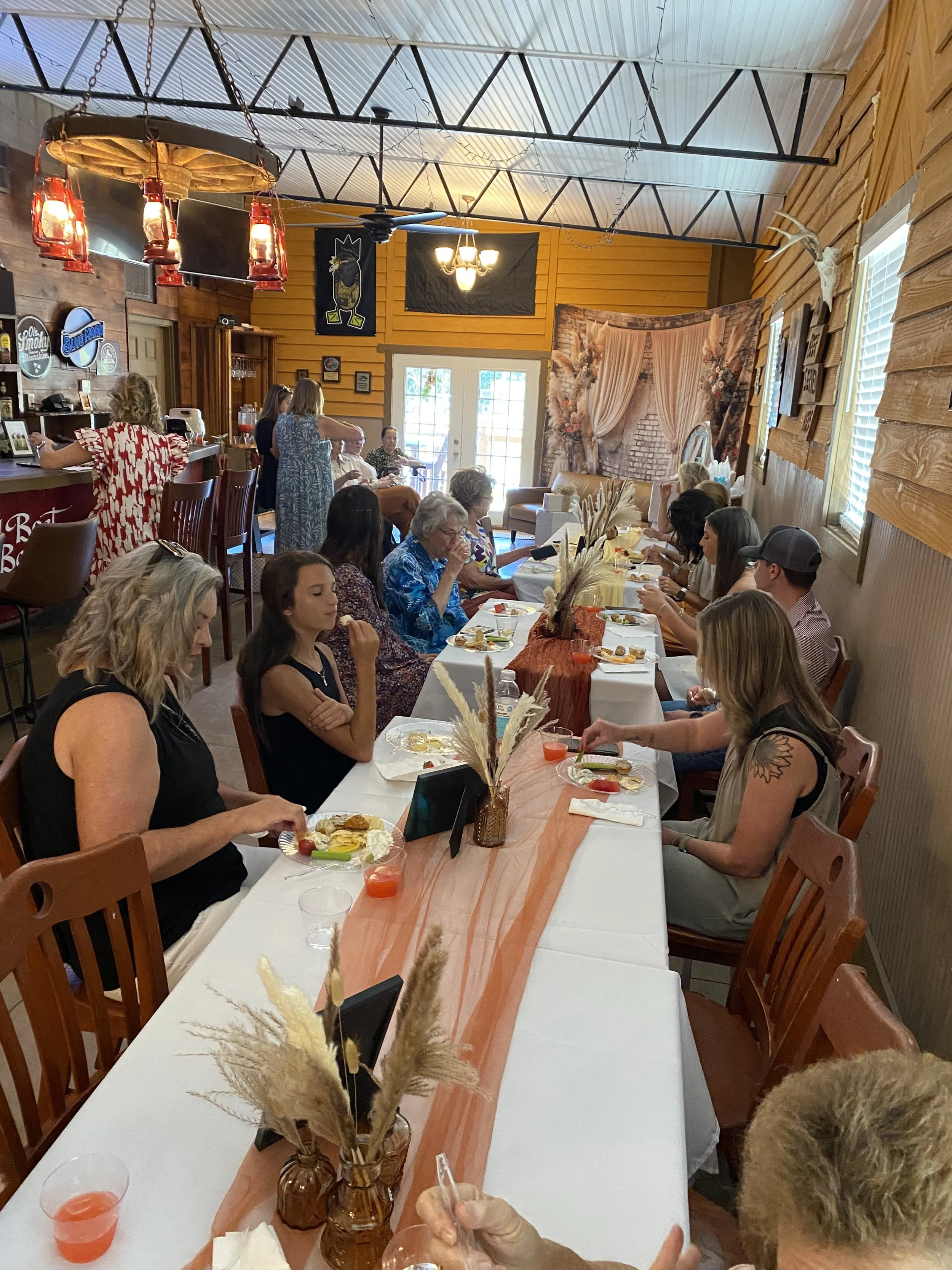 A long dining table with people enjoying a meal in a rustic, decorated restaurant with wood-paneled walls, balloons, and centerpieces featuring dried pampas grass.