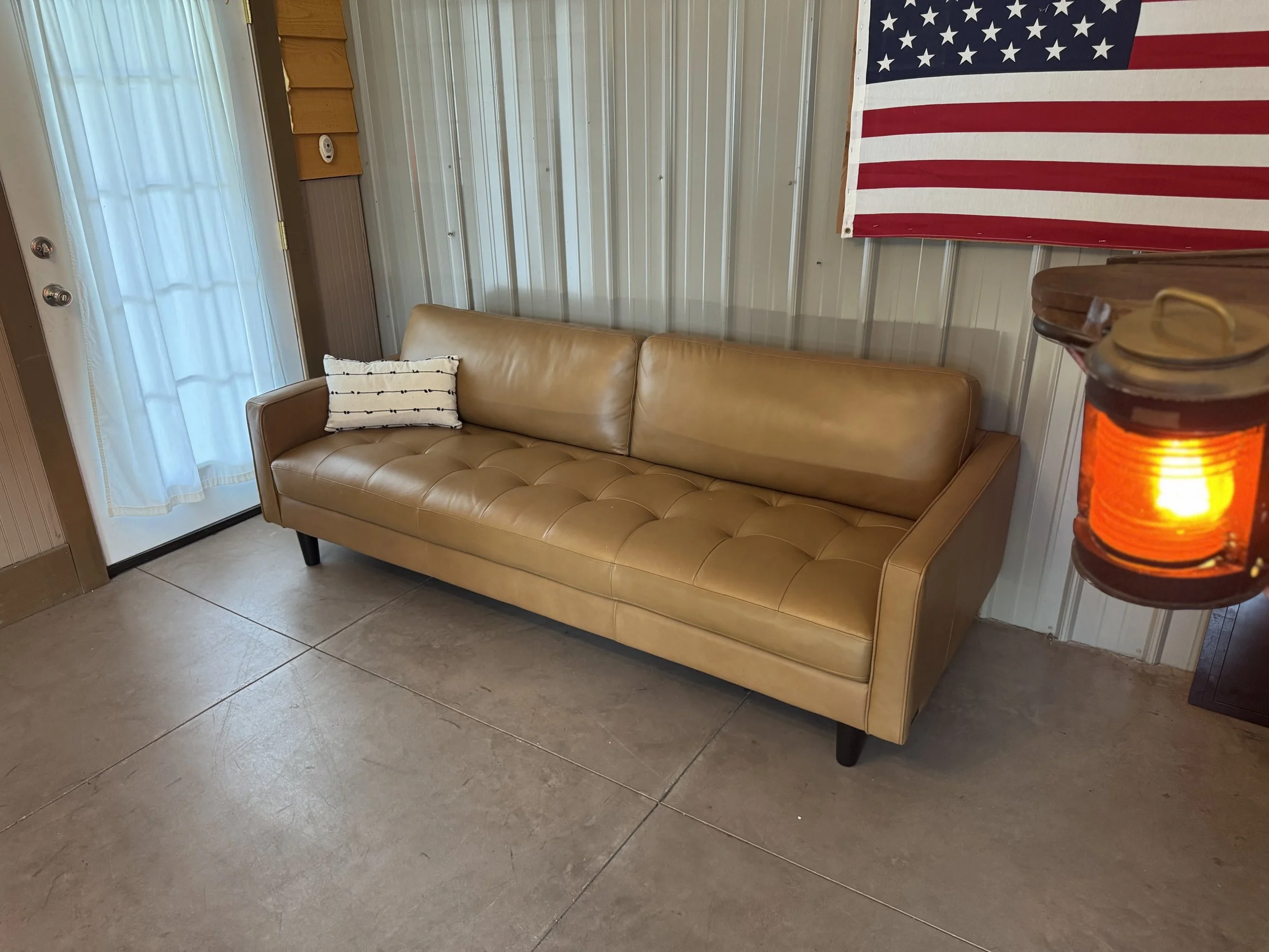 A beige leather couch with a white pillow on the left side, positioned against a wall with metal paneling. To the right of the couch, part of a wooden table and a hanging lantern with an orange glow are visible. Behind the couch, a large American fla