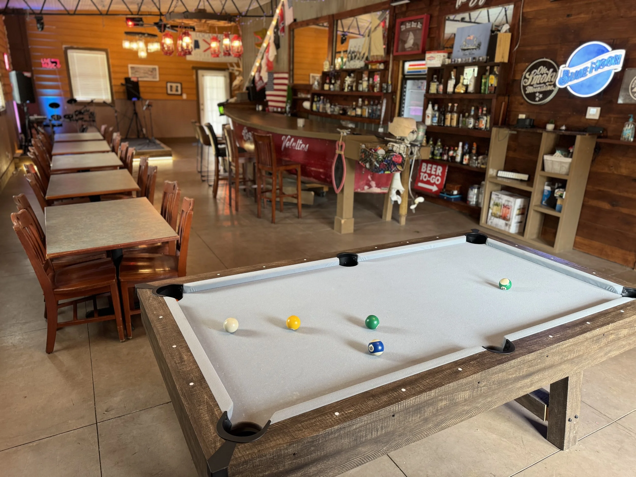 A pool table with four balls on it in a bar or restaurant. The bar has wooden walls, a bar counter with stools, and a shelving unit with bottles. There are tables and chairs along the side, and decorated with signs and artwork.
