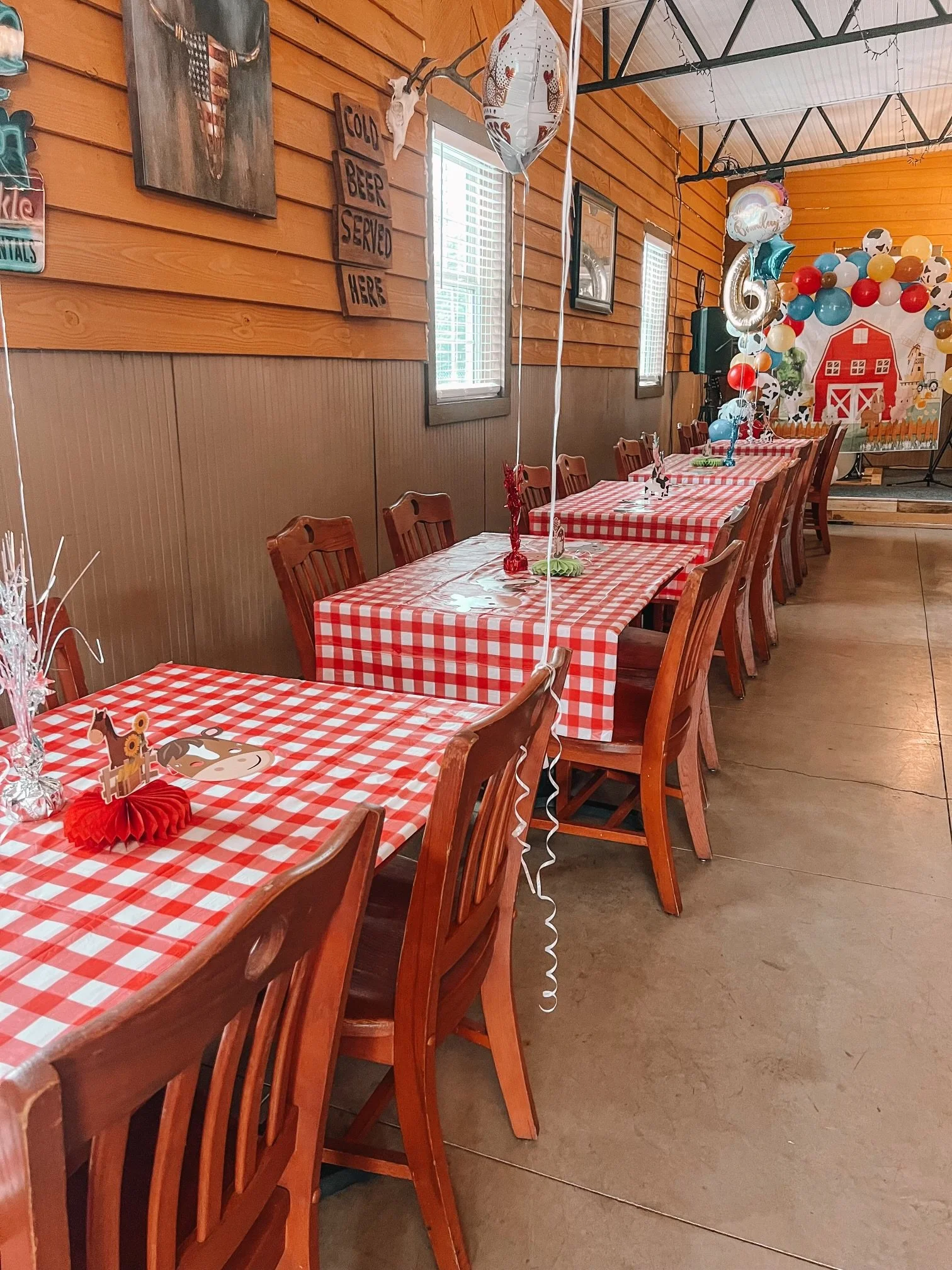 Party tables covered with red and white checkered tablecloths, decorated with balloons, balloons shaped like animals, and paper cut-outs with a farm theme in a rustic indoor setting.