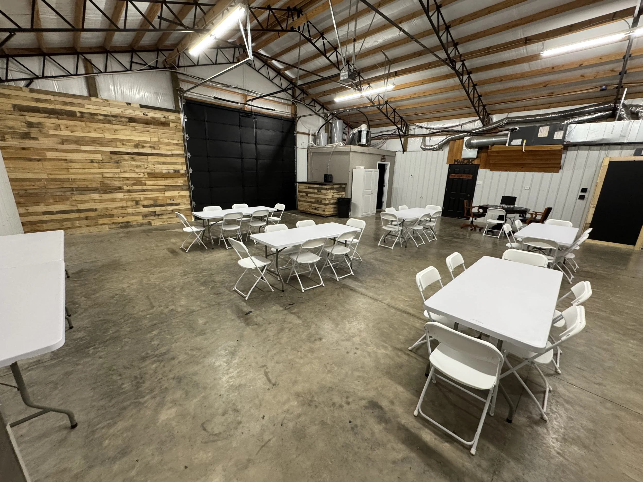 Empty event space with white tables and chairs, wooden wall accents, and a black garage door. Exposed ceiling beams and HVAC ductwork visible.