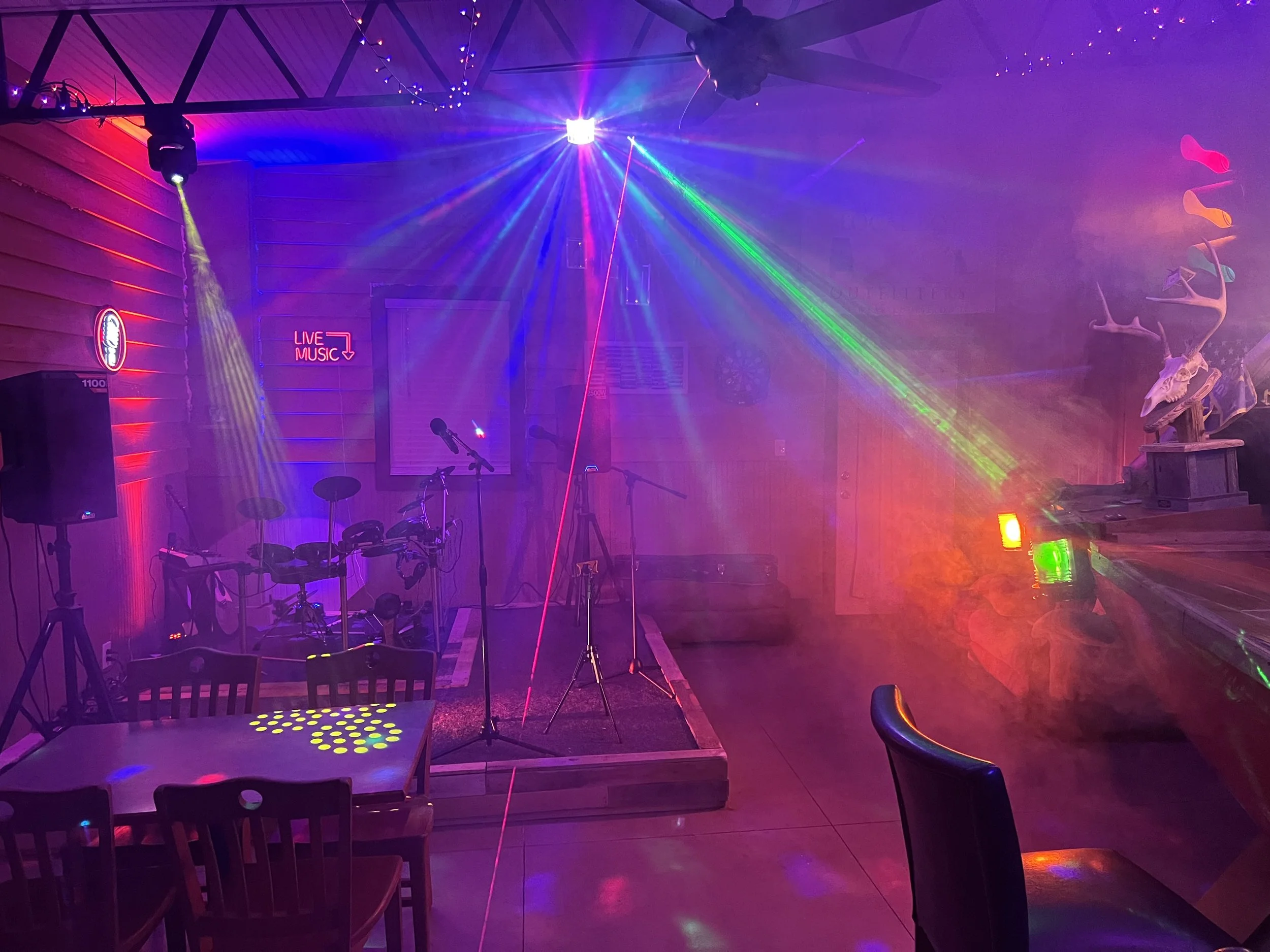 Indoor music venue with colorful laser lights, a drum set on a small stage, a sign that says 'Live Music', and decorative mounted animal skulls with antlers on the right side.