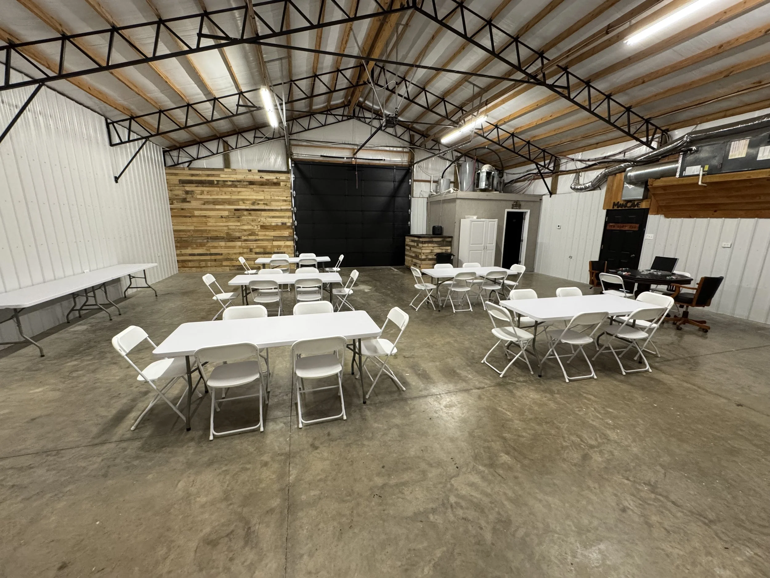 An open industrial-style event space with white folding chairs and tables, a wooden accent wall, and a computer station in the corner.
