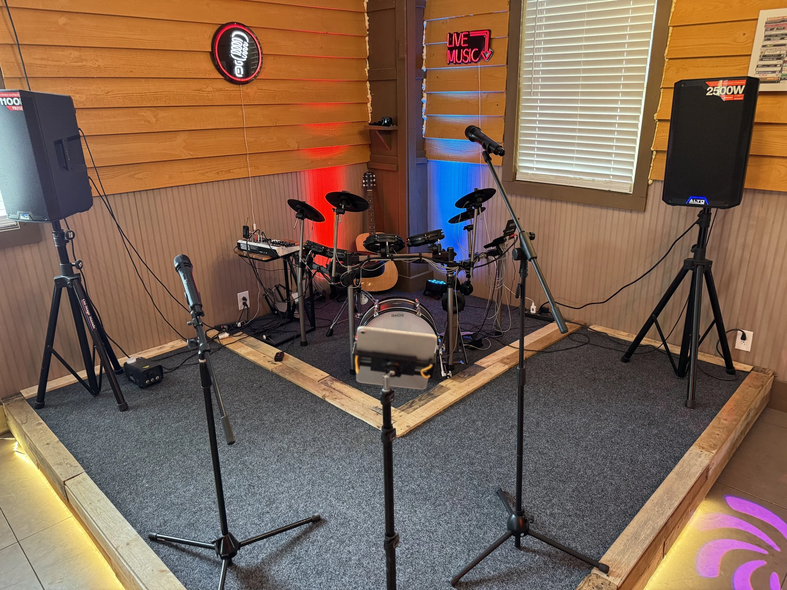 Small stage with electronic drum set, guitar, microphones, and speakers, decorated with red and blue lights, with signs that read 'Live Music' and a musical note.