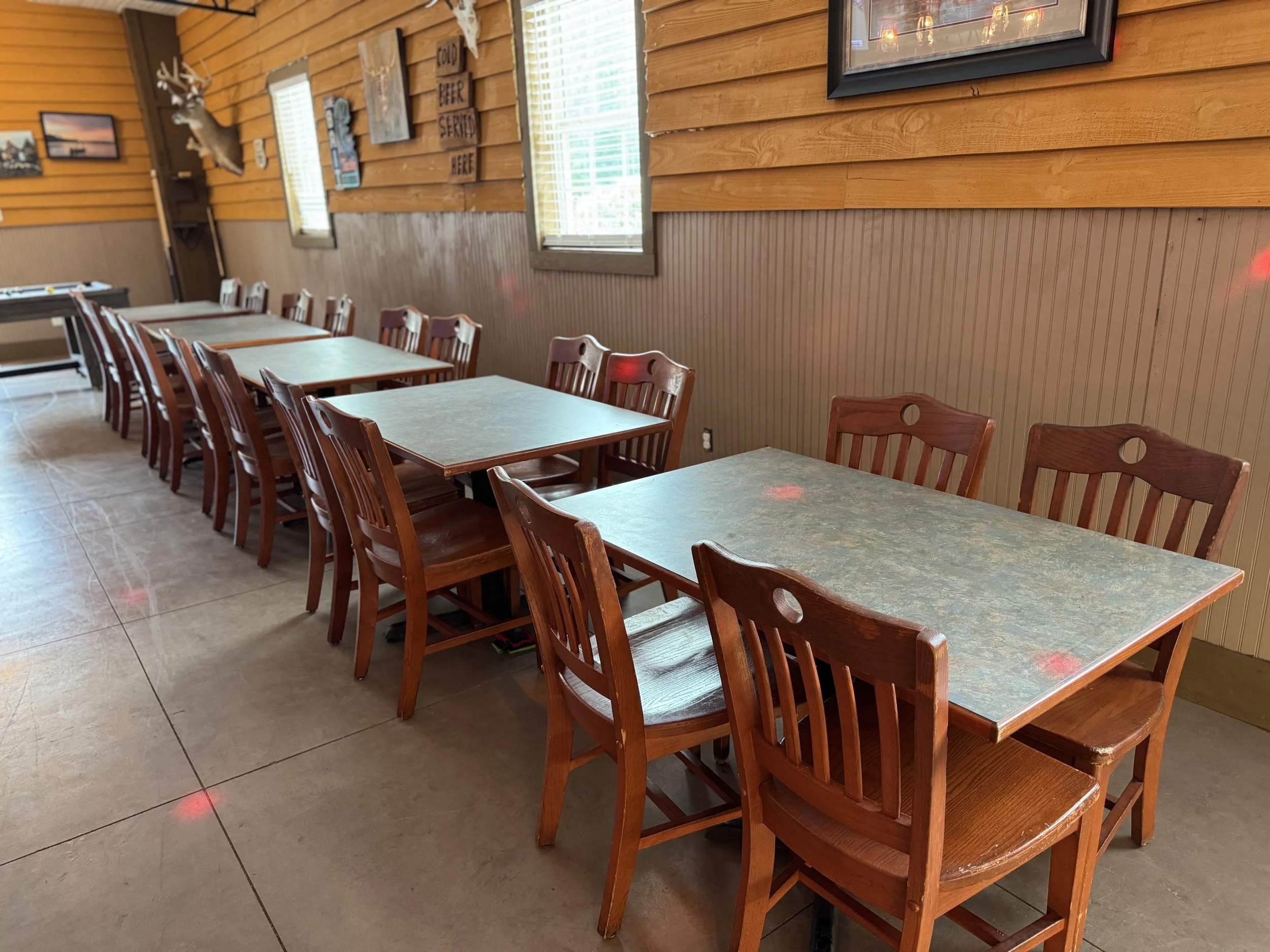 Empty indoor restaurant with wooden tables and chairs arranged in two rows, wood-paneled walls, windows with blinds, and wall decorations including a mounted deer head.