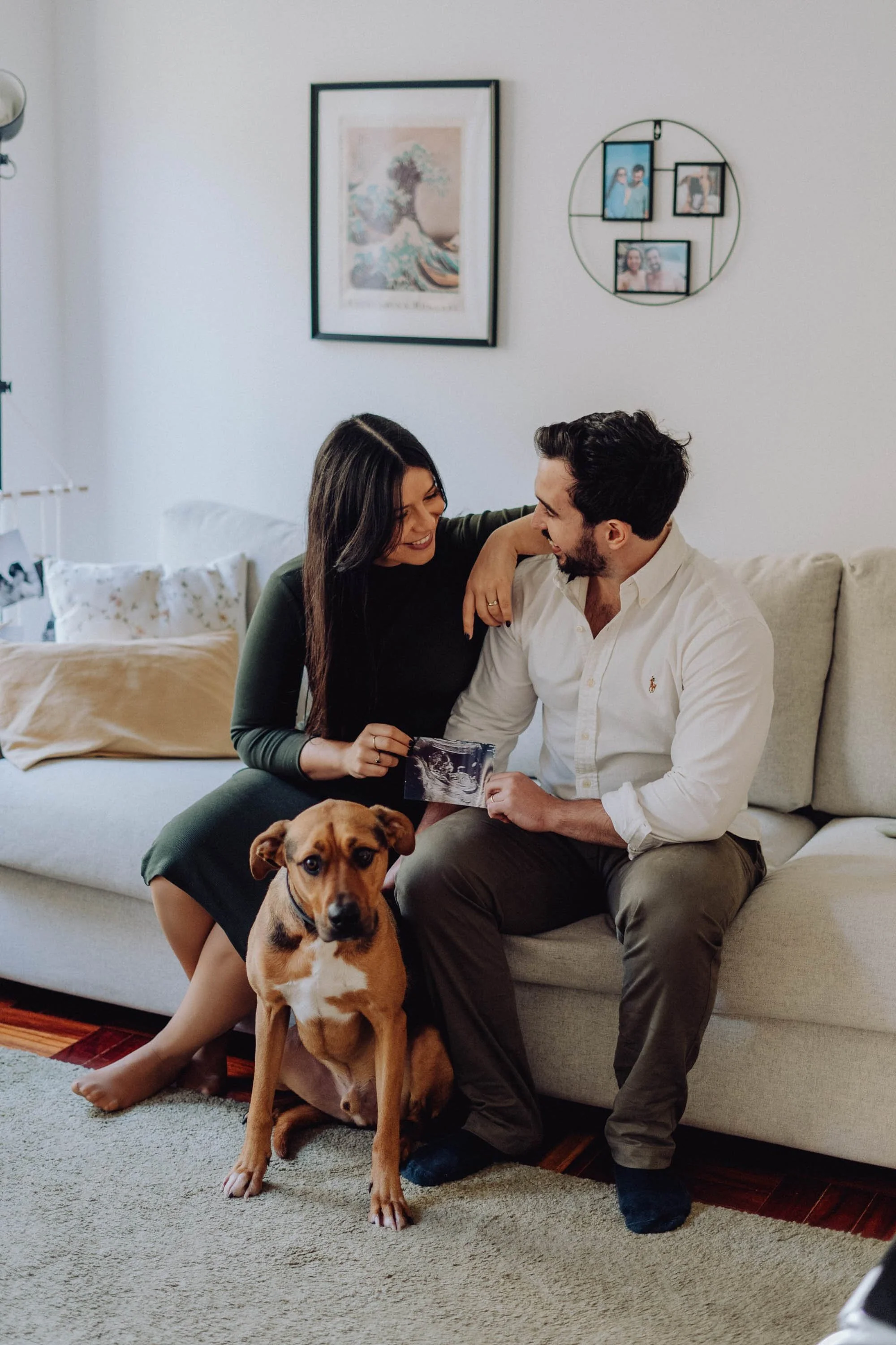 A couple sitting on a beige couch, holding an ultrasound photo, with their dog sitting on the floor. The woman has long dark hair and is wearing a green dress, while the man has dark hair, a beard, and is wearing a white shirt. They are smiling and l