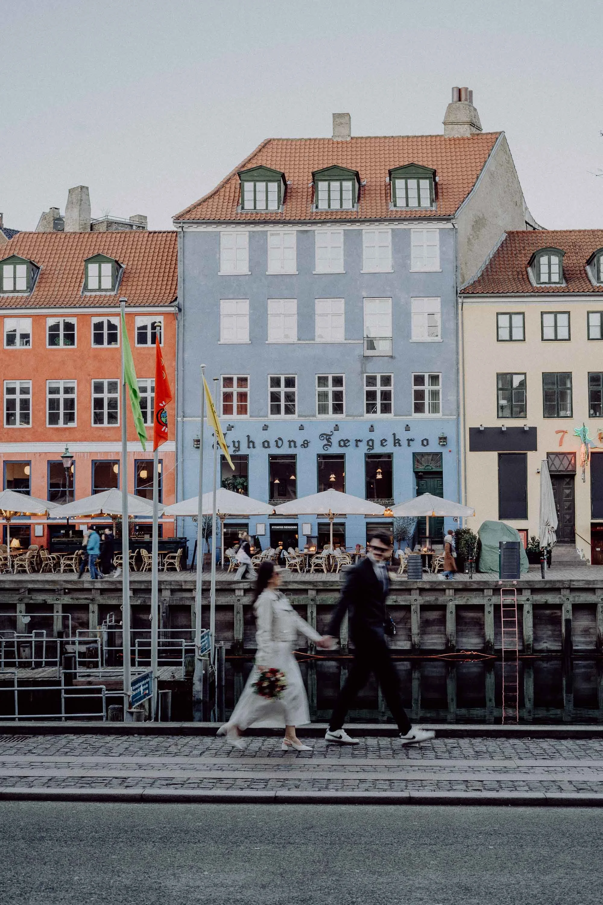 A bride and groom holding hands walking along a cobblestone street in front of colorful buildings and cafes in a European city.