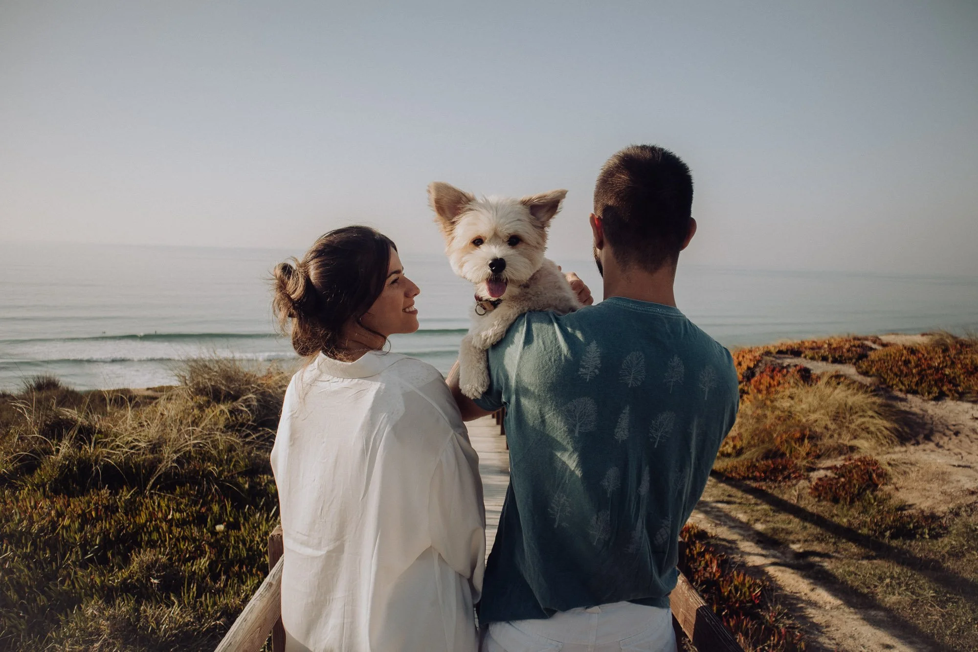 A couple and their dog on a boardwalk overlooking the ocean at sunrise or sunset.
