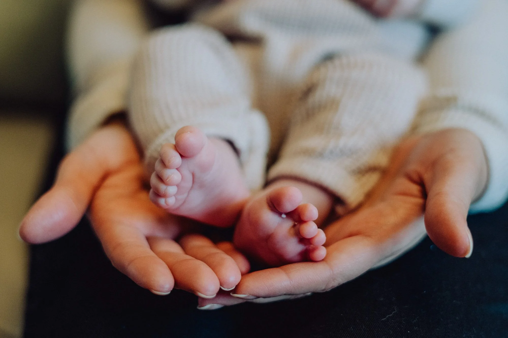 An adult's hands gently holding a small baby's feet, with the baby wearing beige striped pajamas.