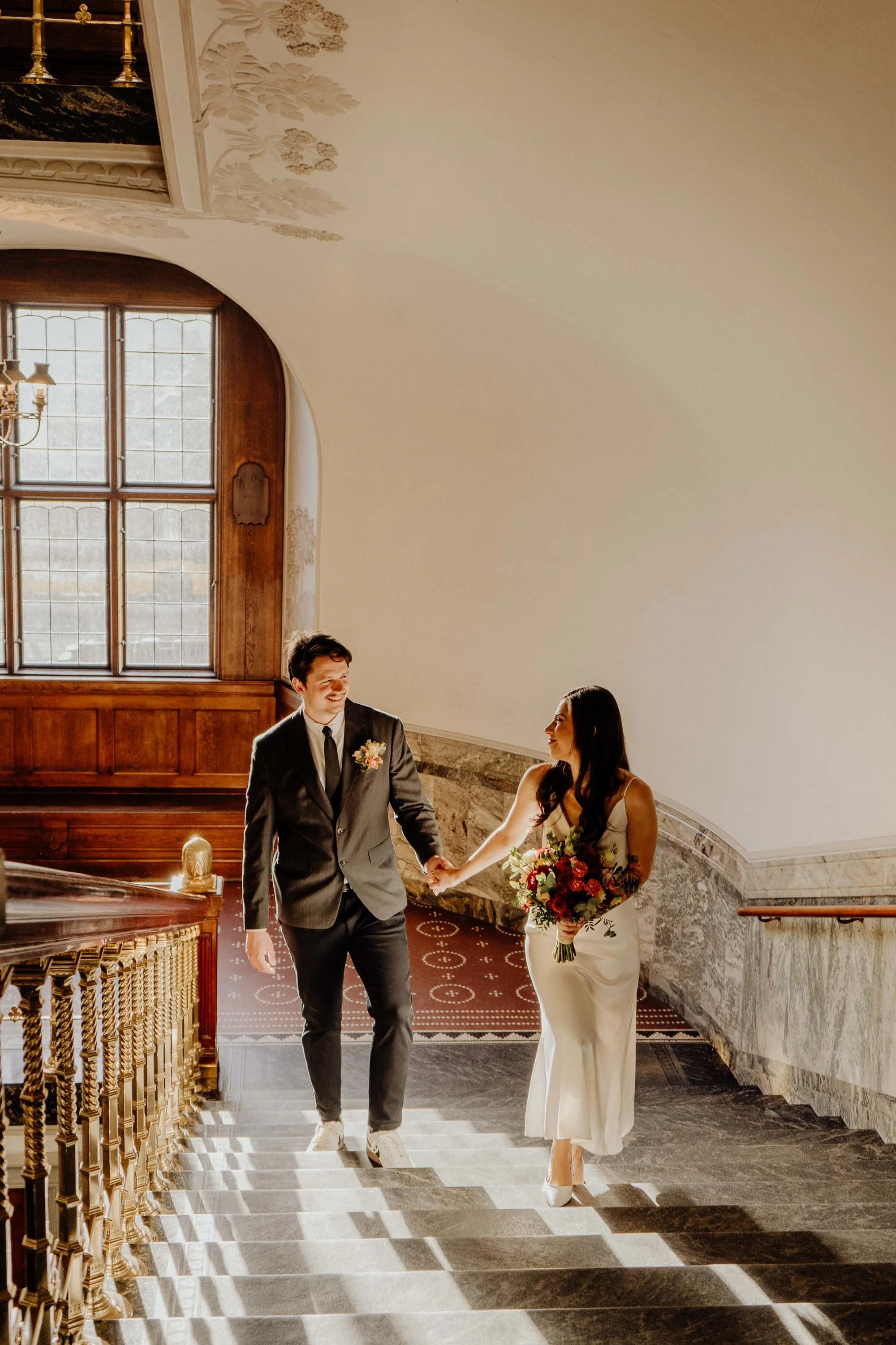 A bride and groom walking up a staircase holding hands, with the bride holding a bouquet of flowers and the groom wearing a suit, in a well-lit, elegant interior space.