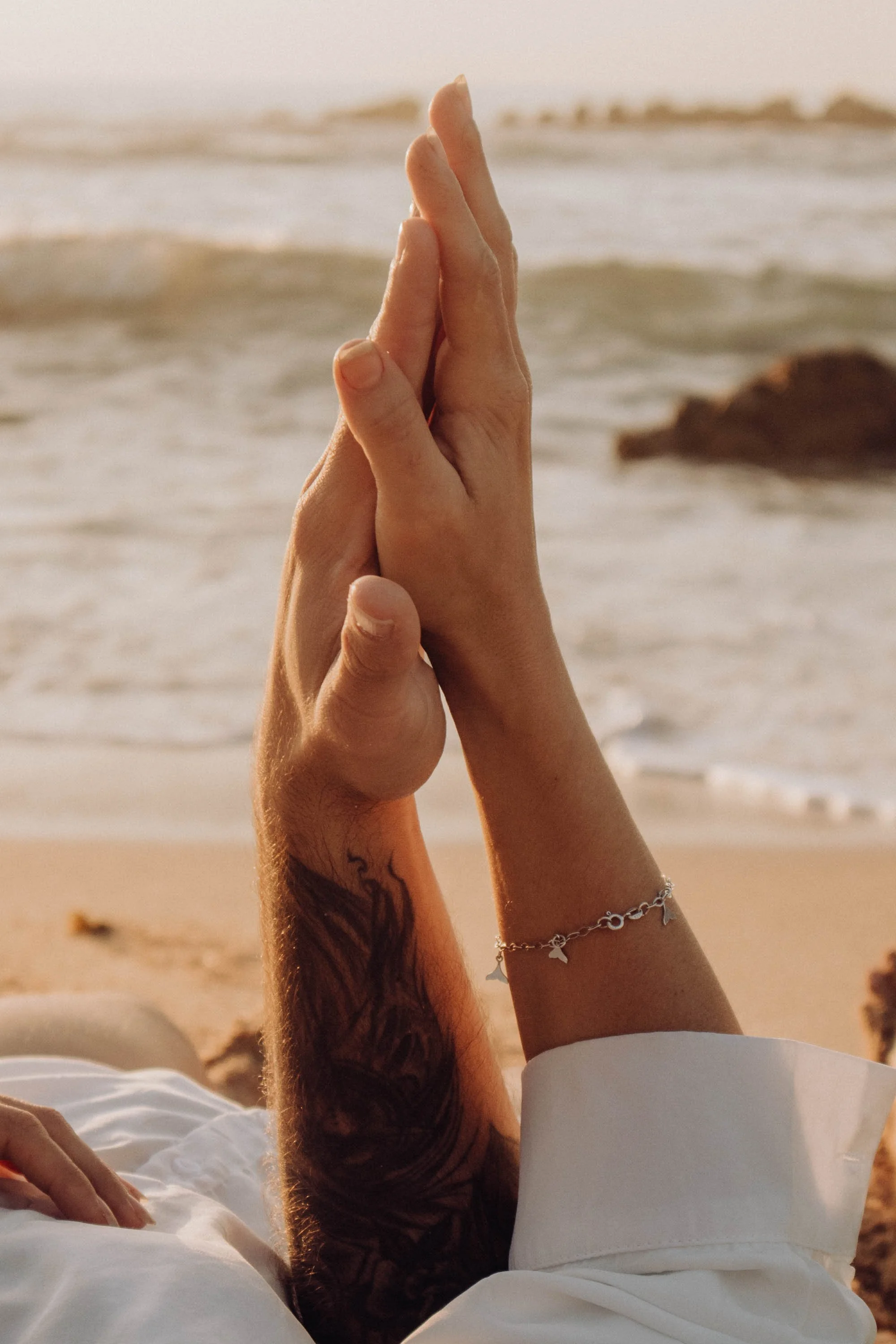 Two hands in a prayer position near a beach with waves and rocks in the background.
