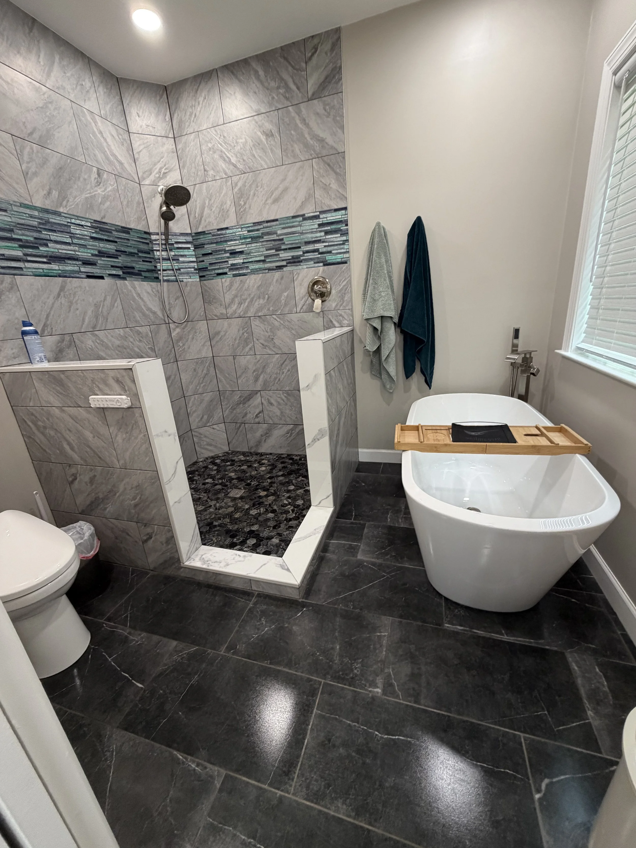 A modern bathroom with dark stone flooring, a walk-in shower with gray marble tiles and a horizontal strip of colorful glass mosaic, a white free-standing bathtub with a wooden tray, two towels hanging on the wall, a window with blinds, and a toilet 