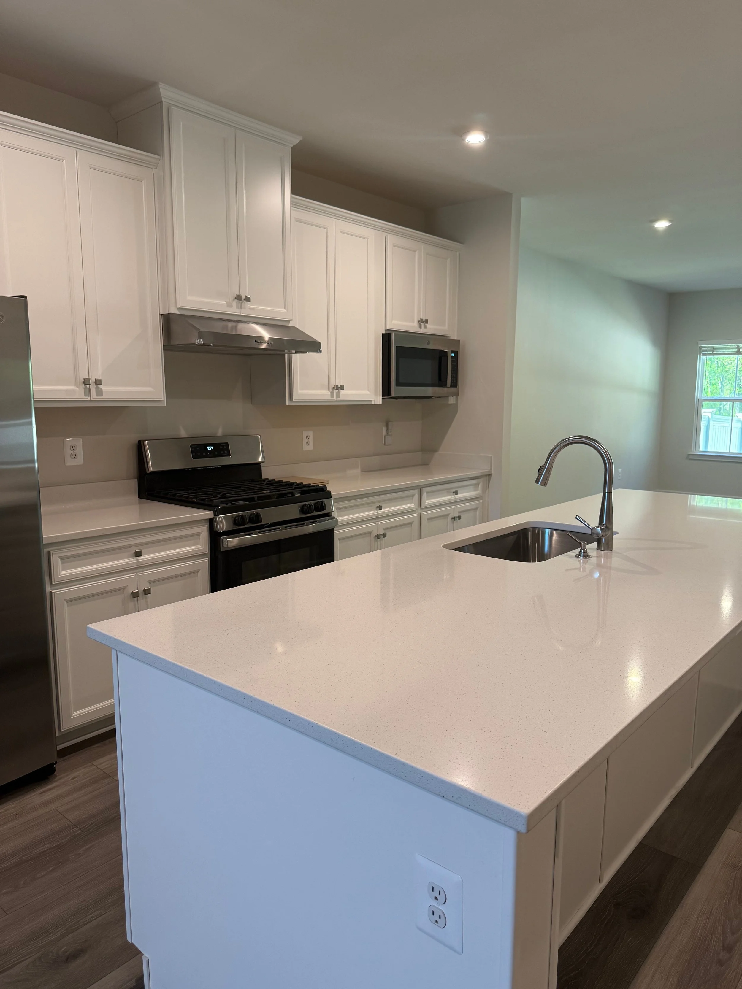 Modern kitchen with white cabinets, a white kitchen island with a built-in sink and faucet, stainless steel stove, microwave, and a partial view of a window in a bright room.