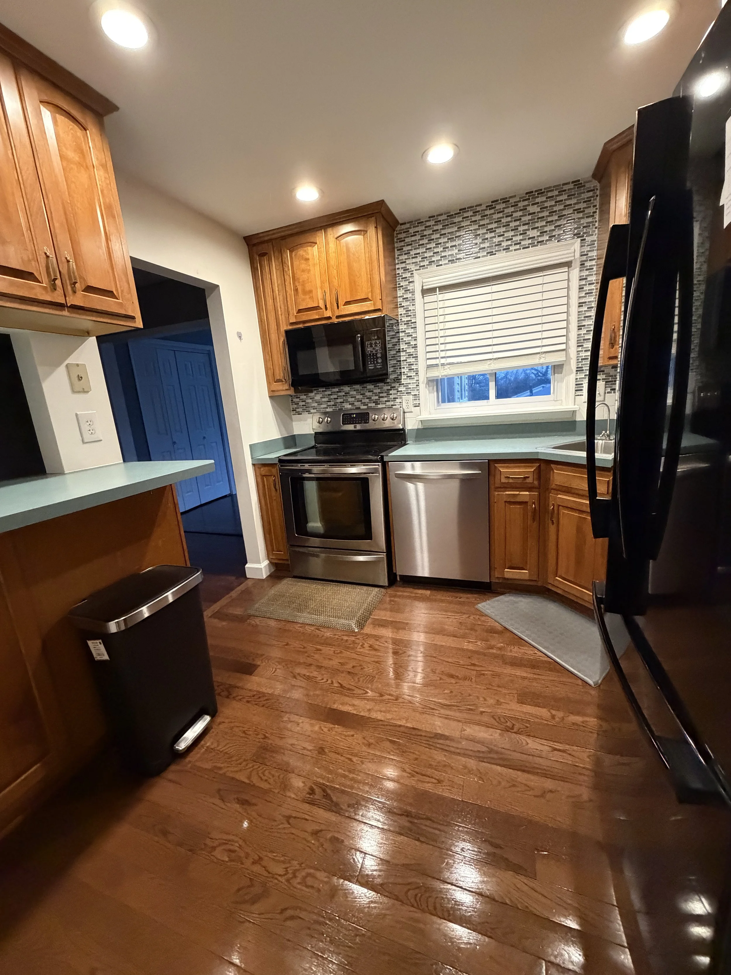 A kitchen with wooden cabinets, stainless steel stove and dishwasher, microwave, black refrigerator, window with blinds, hardwood floors, and recessed lighting.