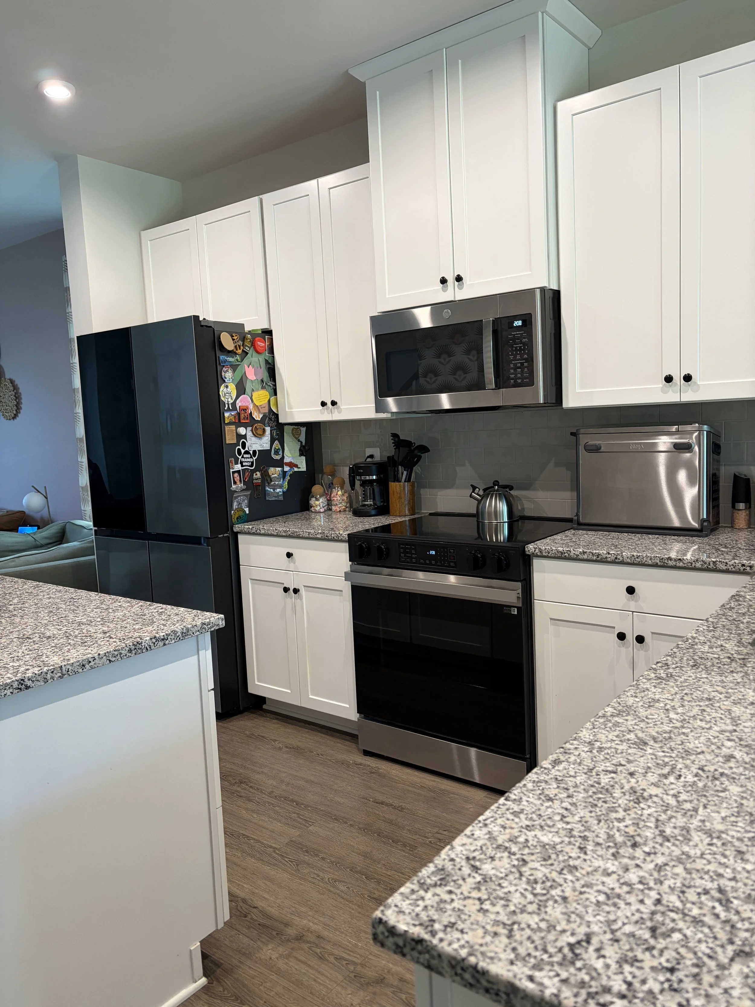 A kitchen with white cabinets, a stainless steel microwave above a black stove, granite countertops, and a black refrigerator decorated with various colorful magnets. The kitchen has wood-look flooring and visible utensils and small appliances on the