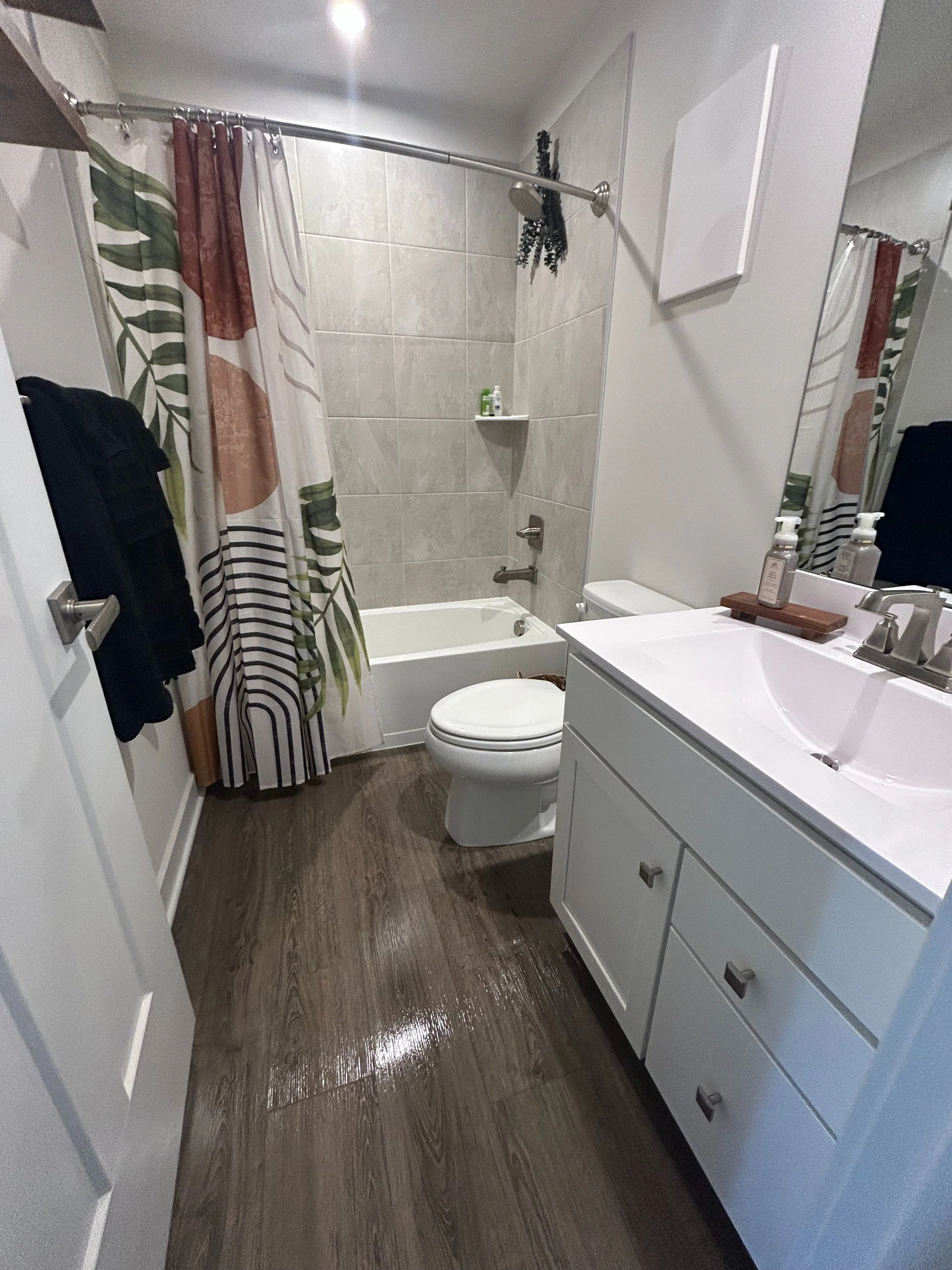 A bathroom with a shower, a toilet, a white vanity, and a mirror. The shower has beige tiles and a curtain with green, brown, and white patterns. There are toiletries on a small shelf in the shower. The floor appears to be wood or wood-look flooring.